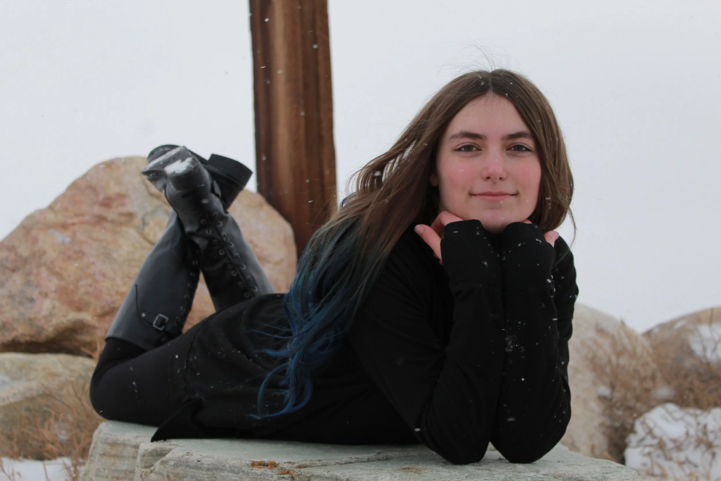 A young woman with long brown hair and blue highlights lying on her stomach on a rock outdoors, smiling at the camera, with snow and rocks in the background.