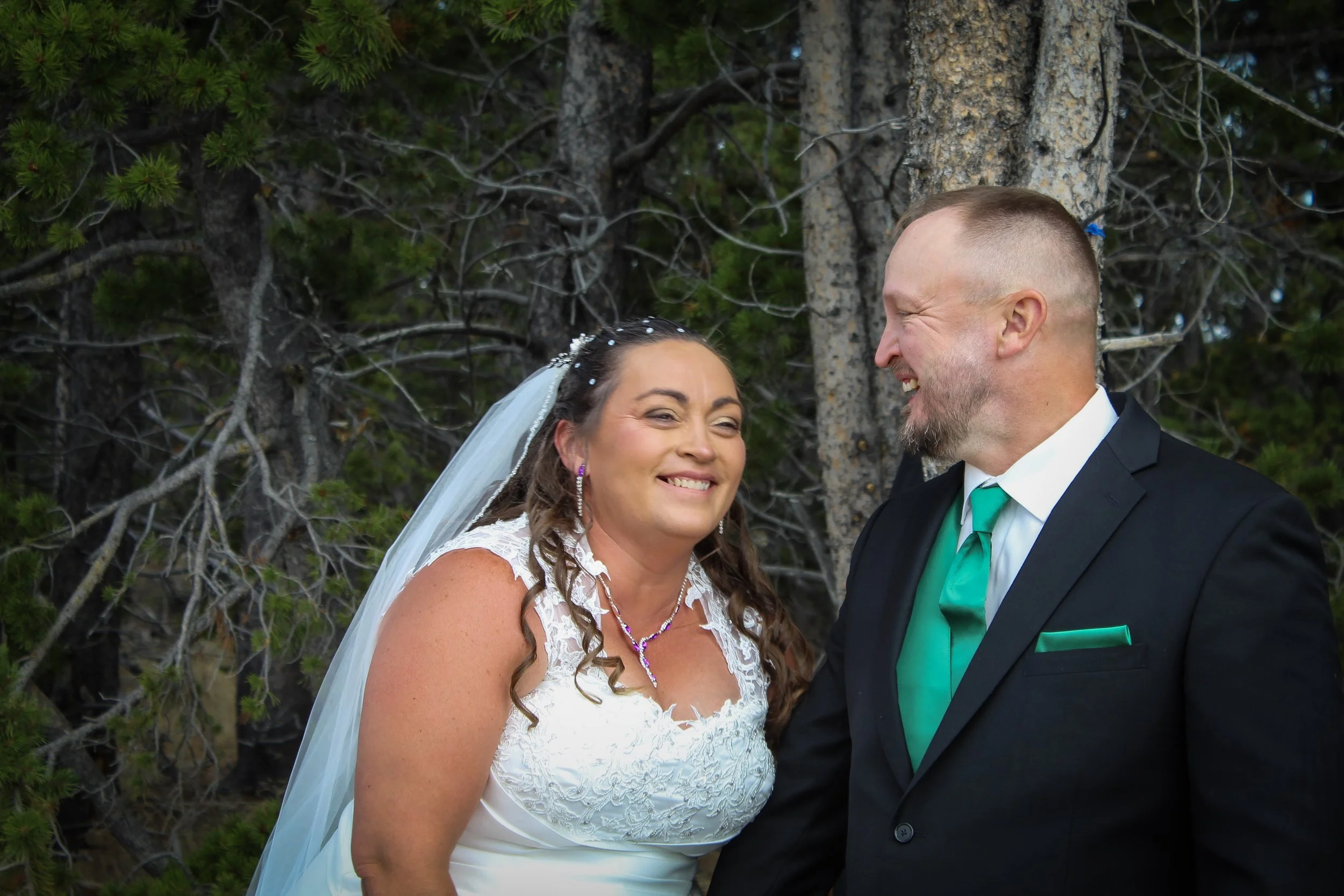 A bride and groom laughing together outdoors near trees, dressed in wedding attire.