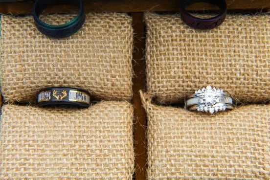 Four rings displayed on burlap-covered stands, including two black rings, a wedding band with a deer design, and a diamond engagement ring with a matching diamond wedding band.