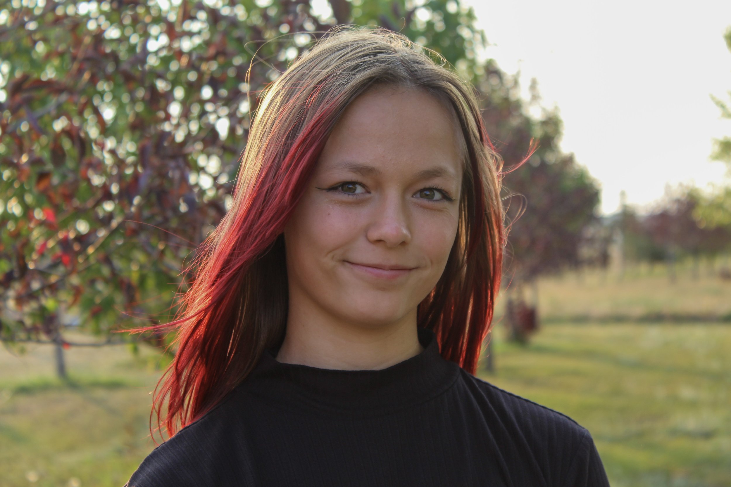 A young woman with brown hair and red highlights standing outdoors in a field with trees in the background, smiling softly at the camera during daylight.