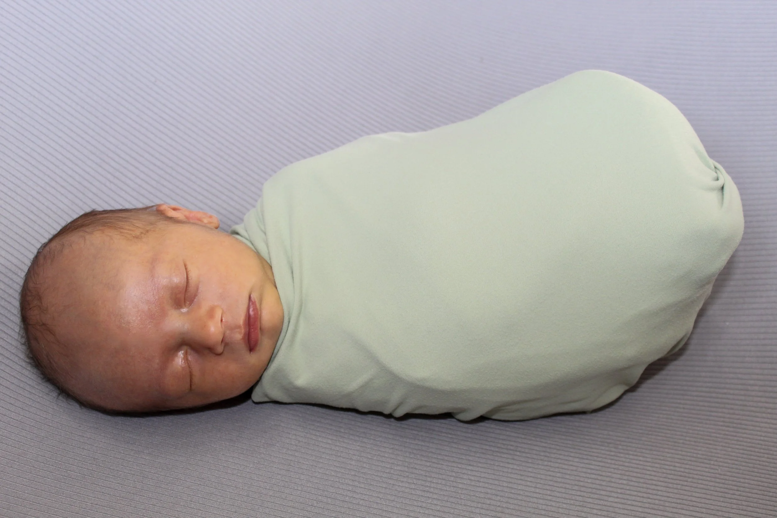 A newborn baby wrapped in a light green swaddle blanket, sleeping on a gray surface.