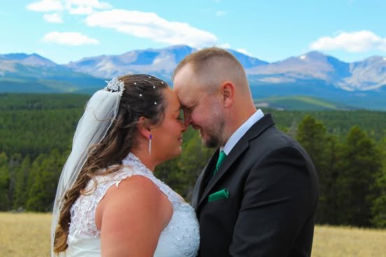 Bride and groom with foreheads touching in a field with mountains and trees in the background.