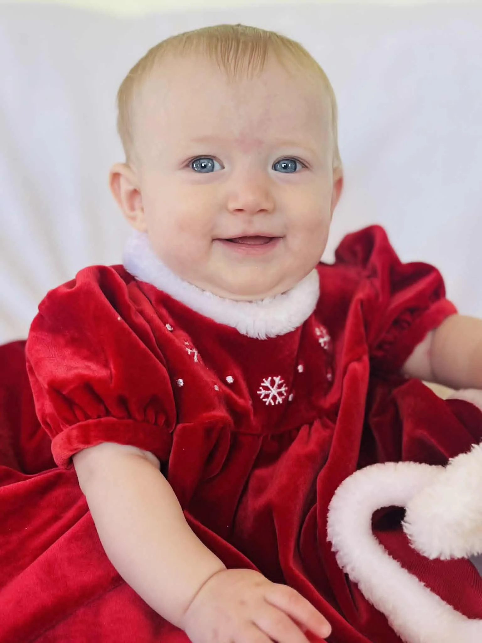 A smiling baby dressed in a red velvet Christmas dress with white snowflake embroidery and a white collar, sitting on a white surface.