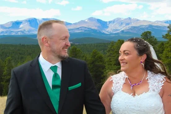 A smiling couple during a wedding ceremony outdoors with mountains and trees in the background.