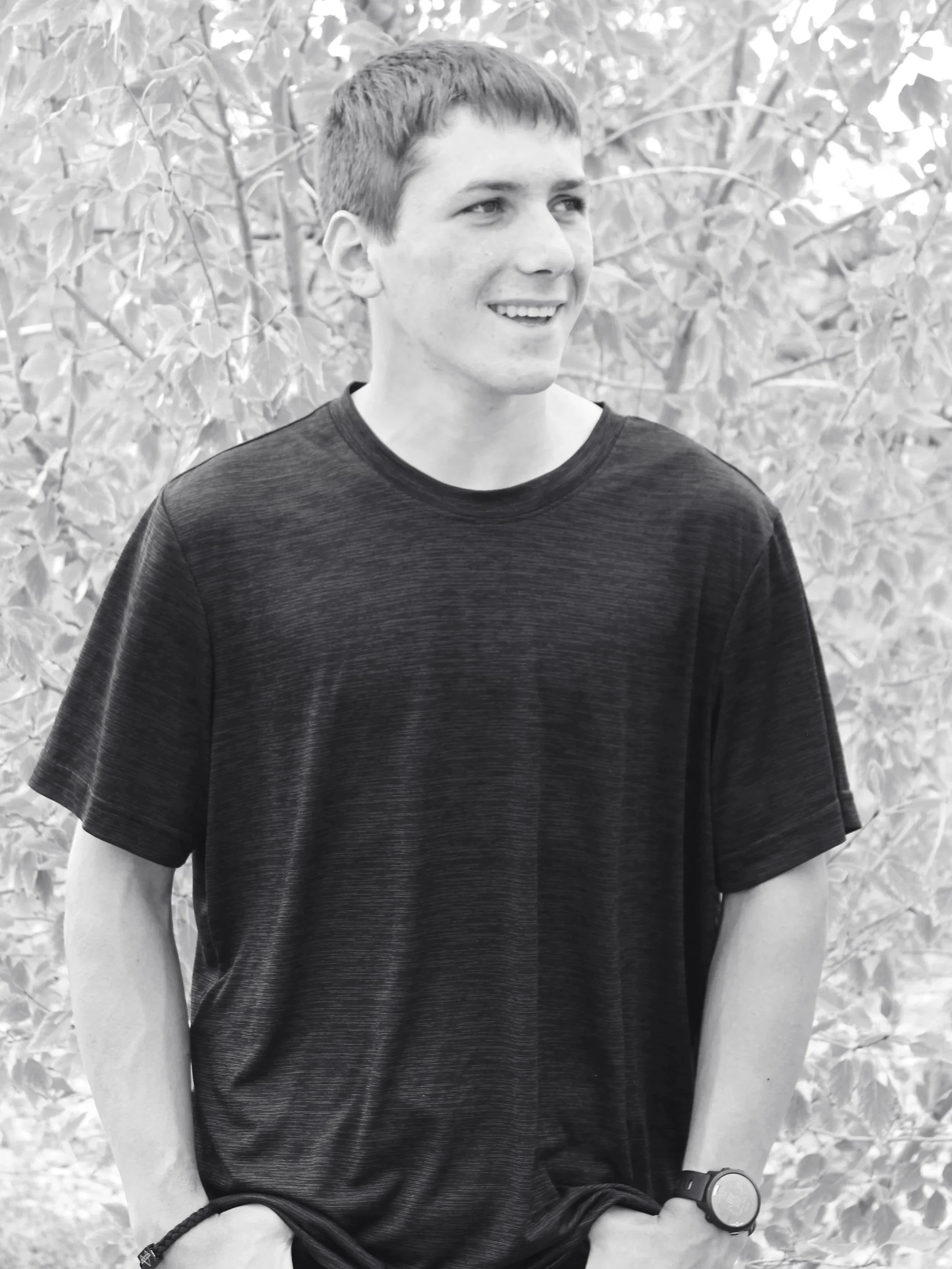 A young man with short hair, wearing a dark t-shirt, stands outdoors in front of leafy trees, smiling and looking to the side.