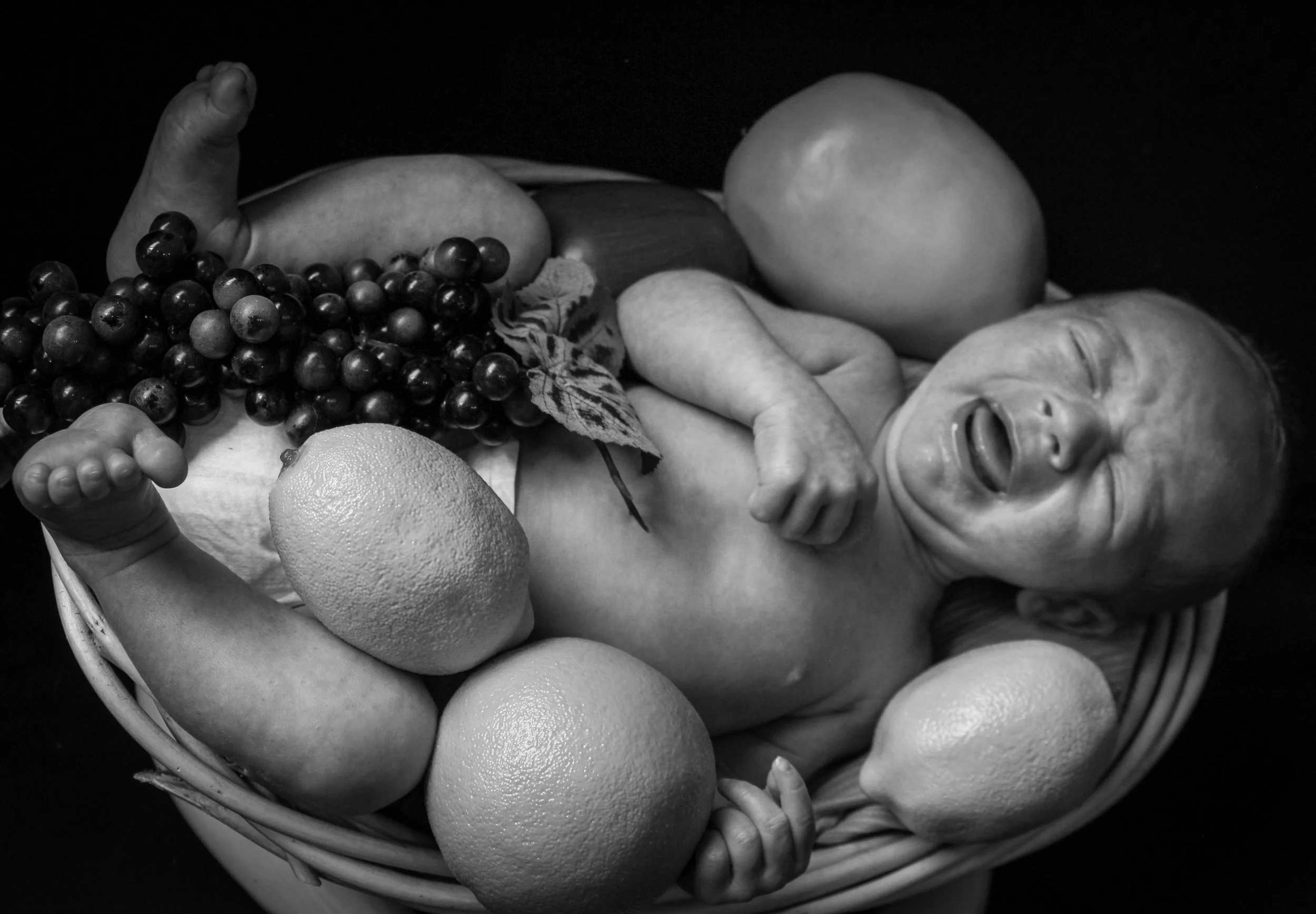 Black and white photo of a newborn baby crying in a basket surrounded by fruits including grapes, oranges, and apples.