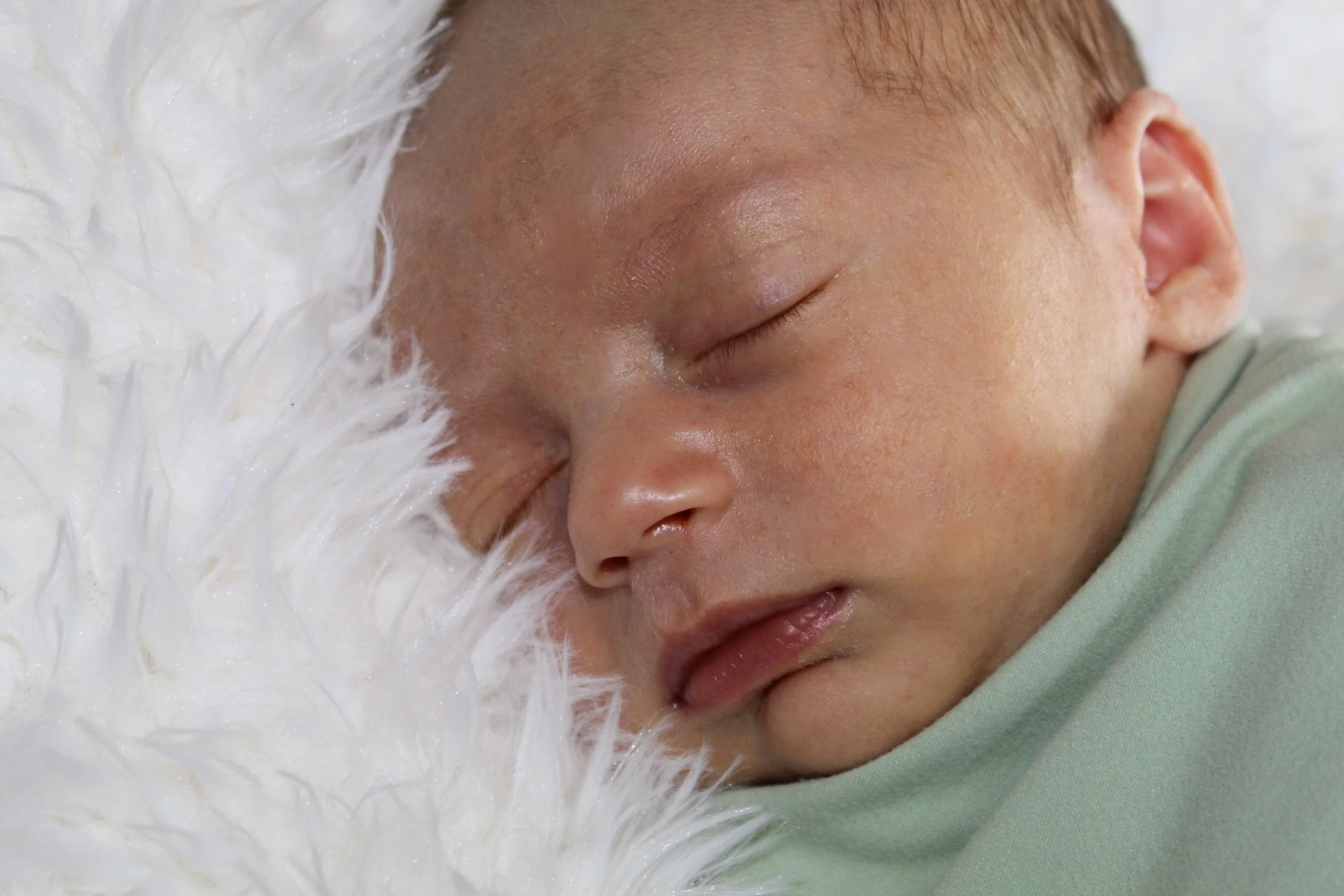 Close-up of a sleeping baby with closed eyes, lying on a soft white furry surface, wearing a light green outfit.