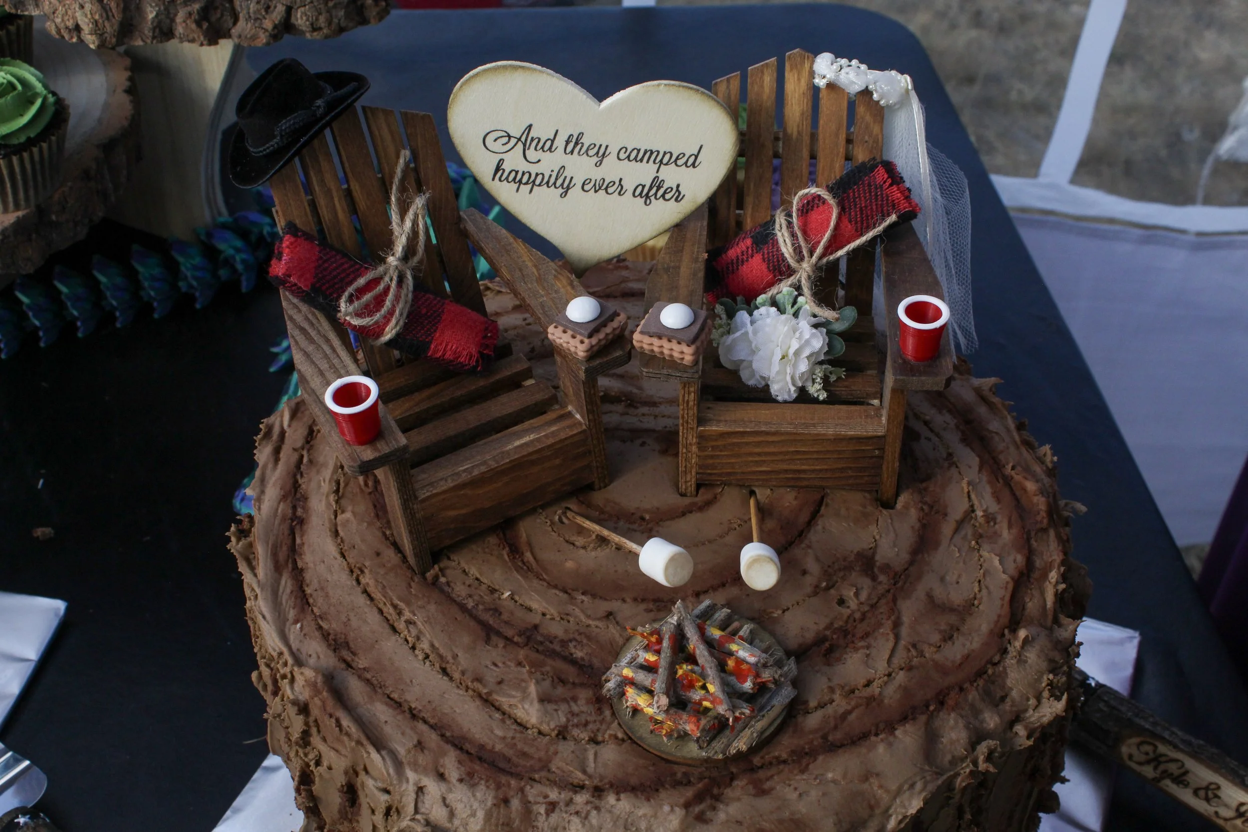 A cake decorated with a miniature wedding scene, including two wooden chairs, small props like wine glasses, marshmallows on sticks, and a heart-shaped sign that reads 'And they camped happily ever after.'