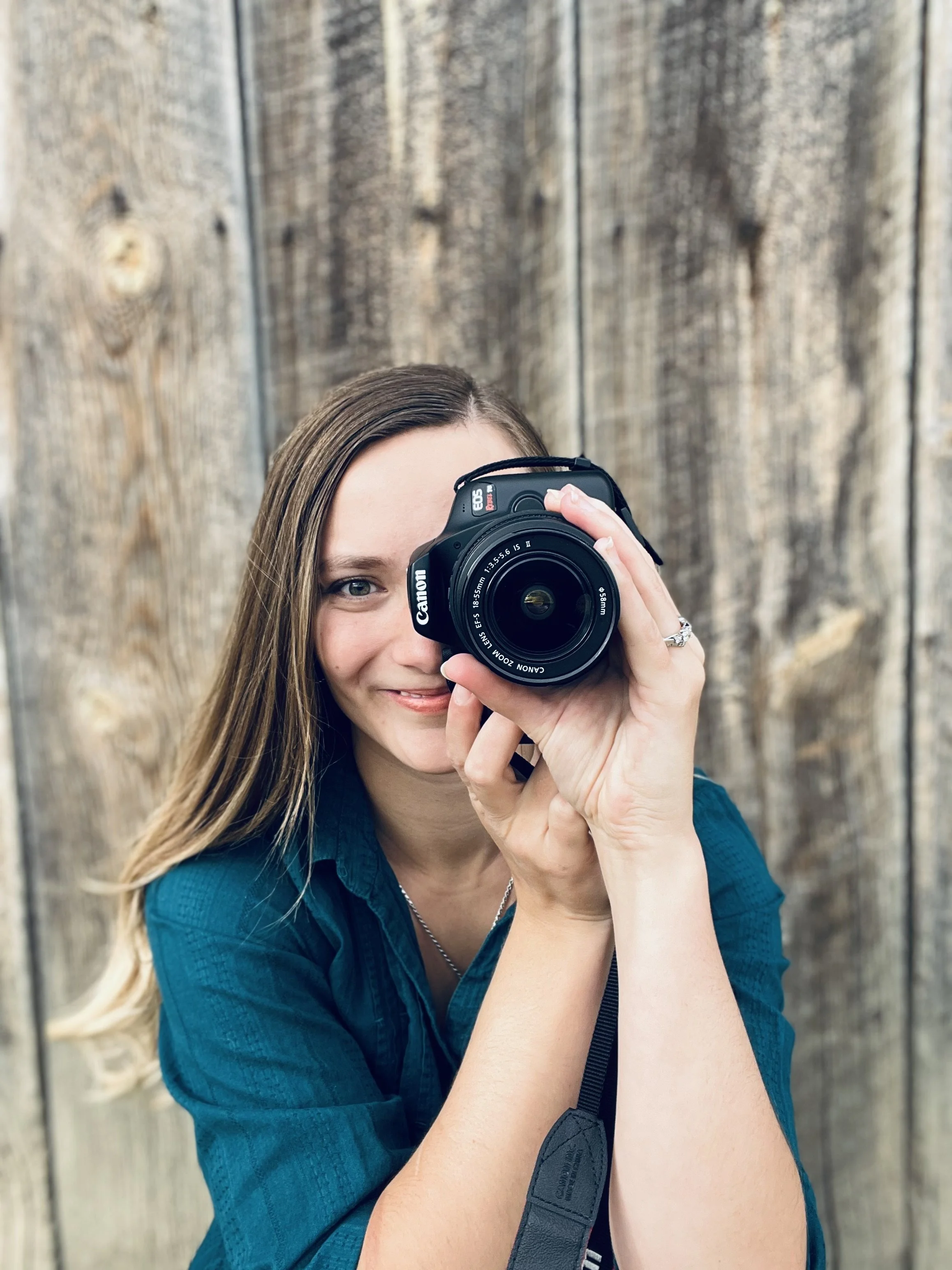 A woman with long brown hair holding a Canon DSLR camera, smiling in front of a wooden fence.