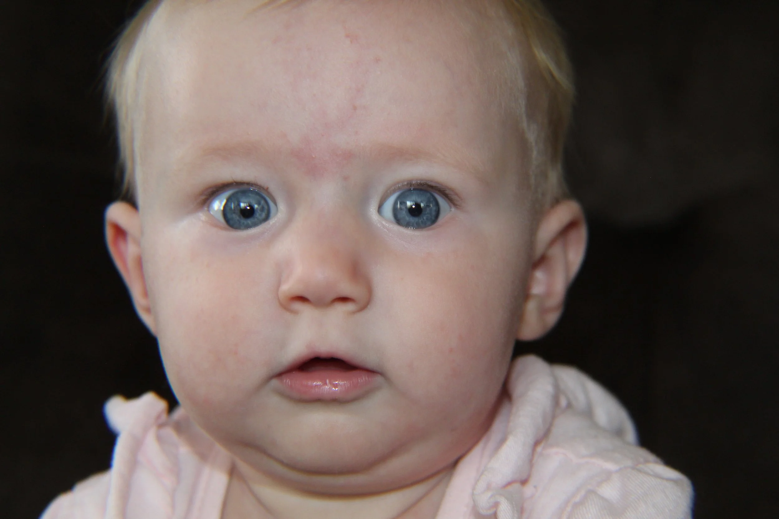 Close-up of a baby girl with bright blue eyes and light-colored hair, wearing a pink outfit, against a dark background.
