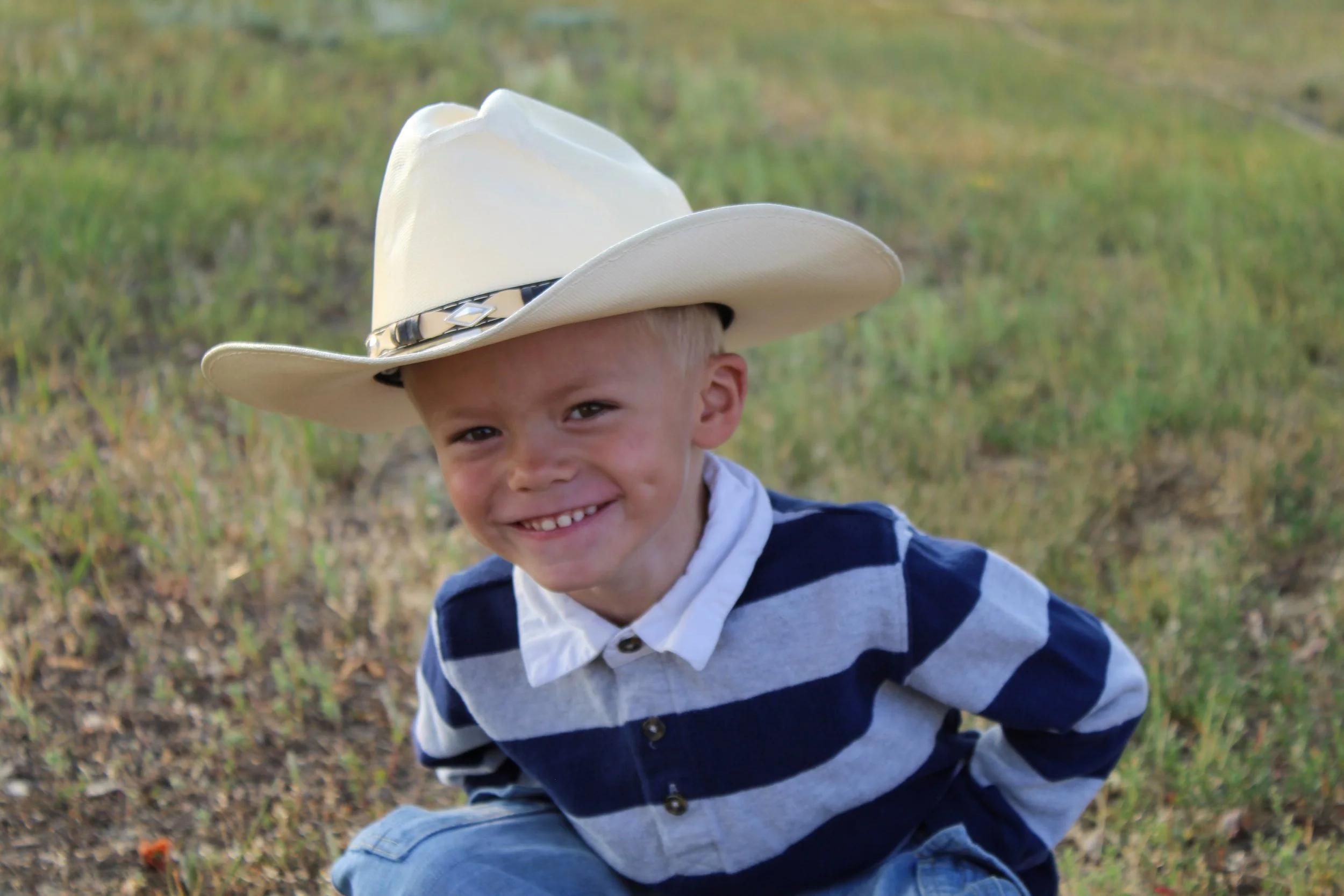 A young boy smiling outdoors wearing a large white cowboy hat and a striped blue and gray shirt, with a grassy field in the background.