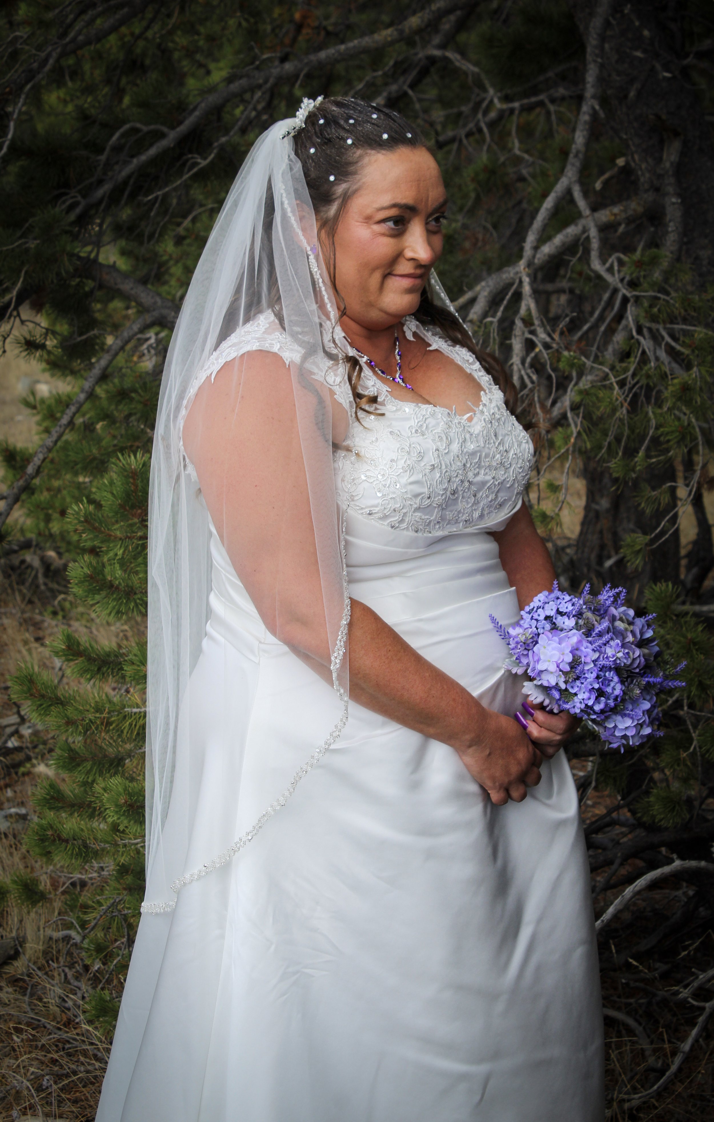 A woman in a white wedding dress holding a bouquet of purple flowers, standing outdoors in front of trees.