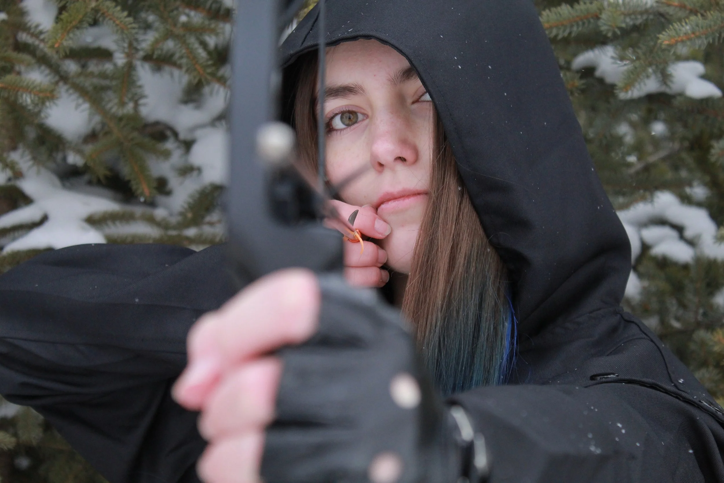 A young woman with long brown hair, wearing a black hoodie, is in a snowy outdoor setting, pointing a bow towards the camera, with snow-covered pine trees in the background.