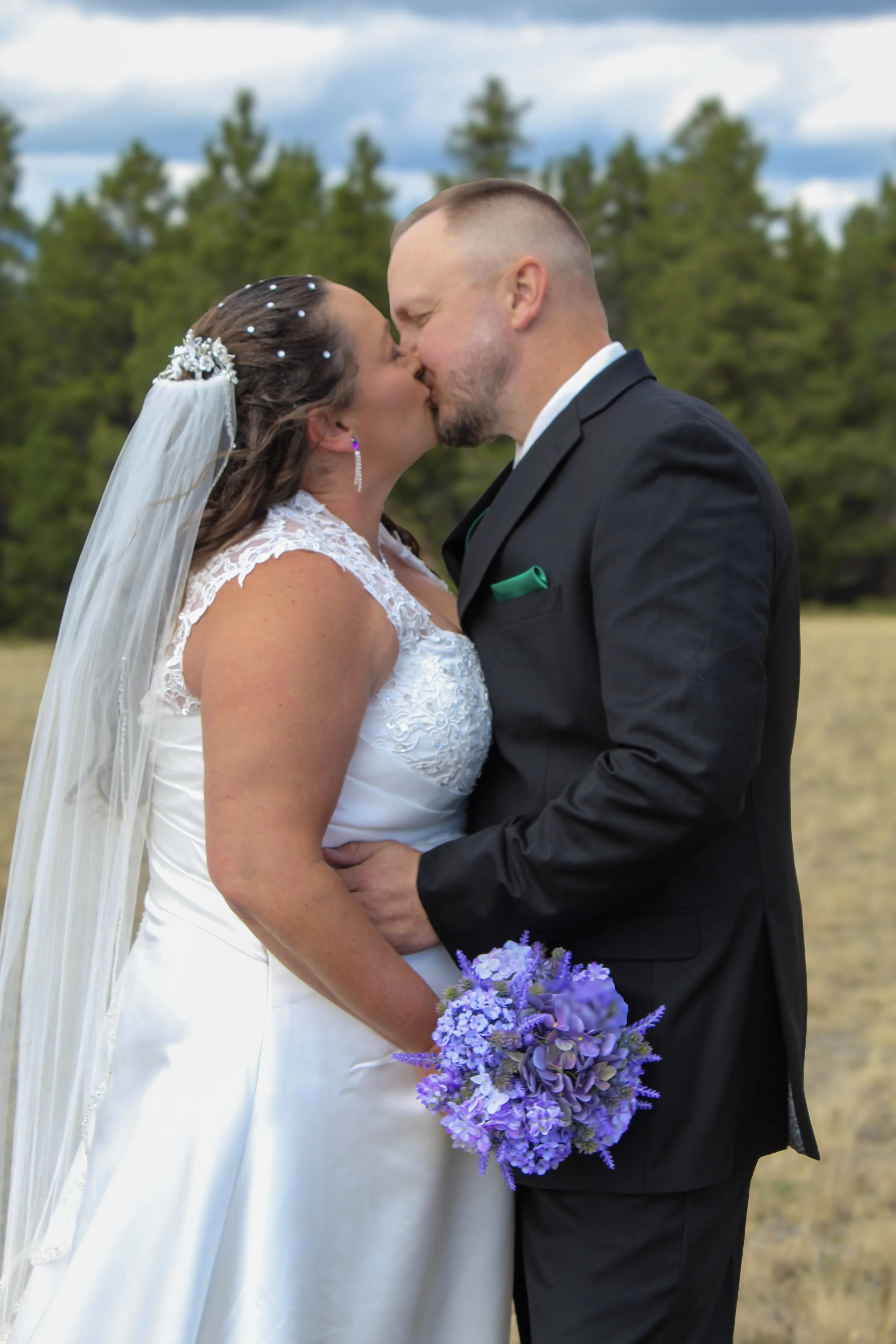 A bride and groom sharing a kiss outdoors, with trees and cloudy sky in the background. The bride is wearing a white wedding dress with lace detail, holding a purple bouquet, and has a veil and jewelry. The groom is in a black suit with a green pocket square.