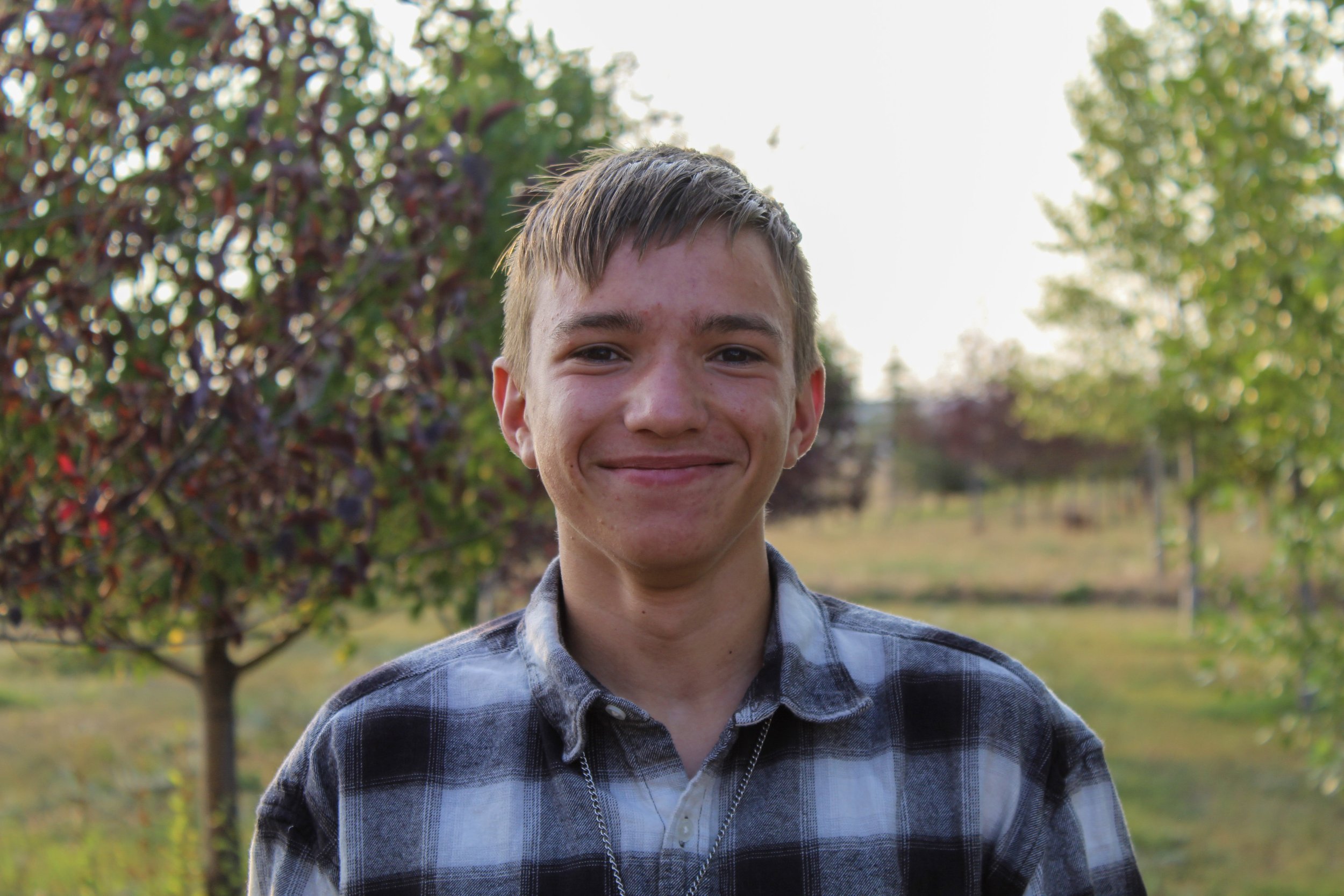 A young man with short brown hair, smiling, stands outdoors with trees and greenery in the background.