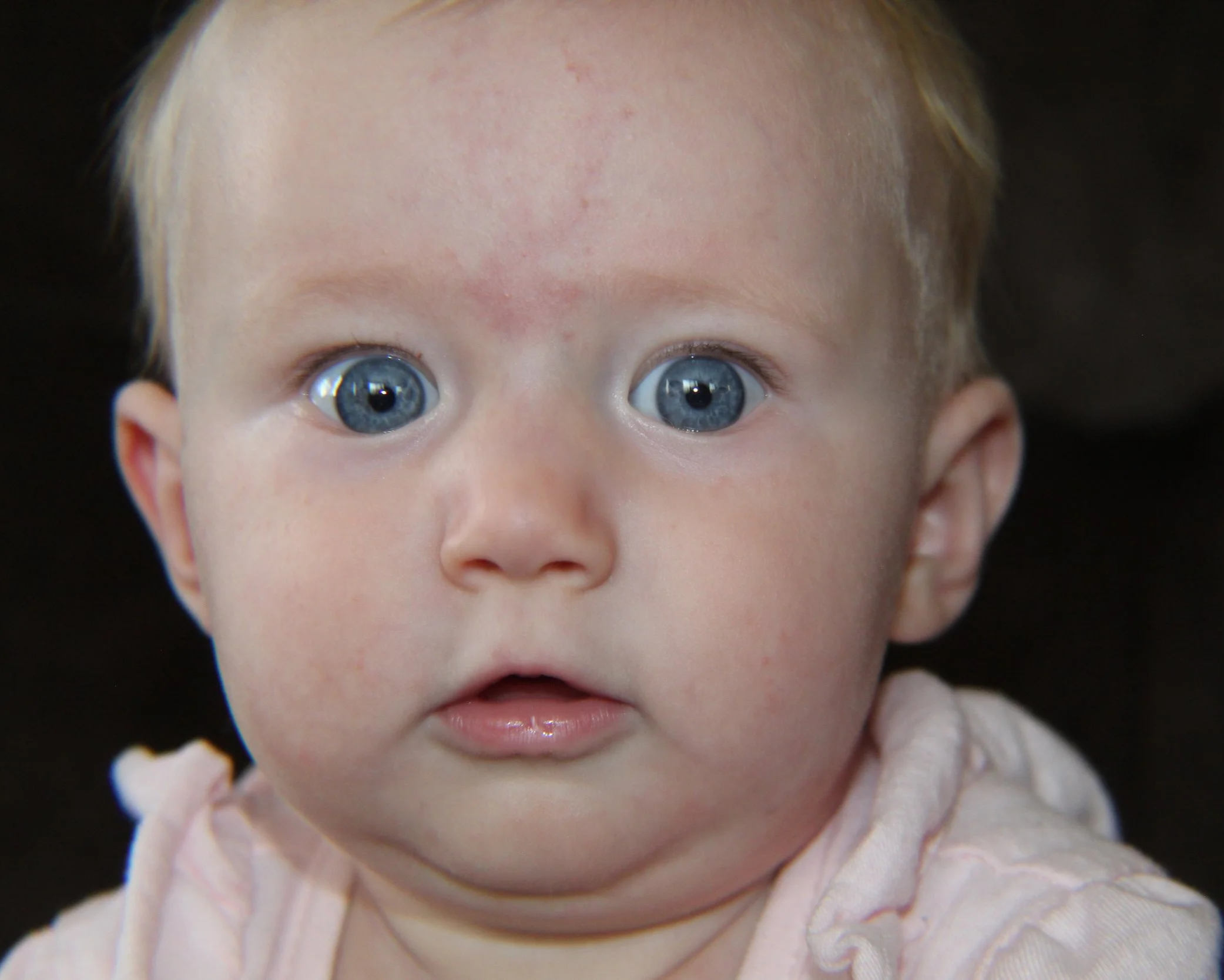 Close-up of a baby with big blue eyes and light-colored hair, wearing a pale pink outfit.