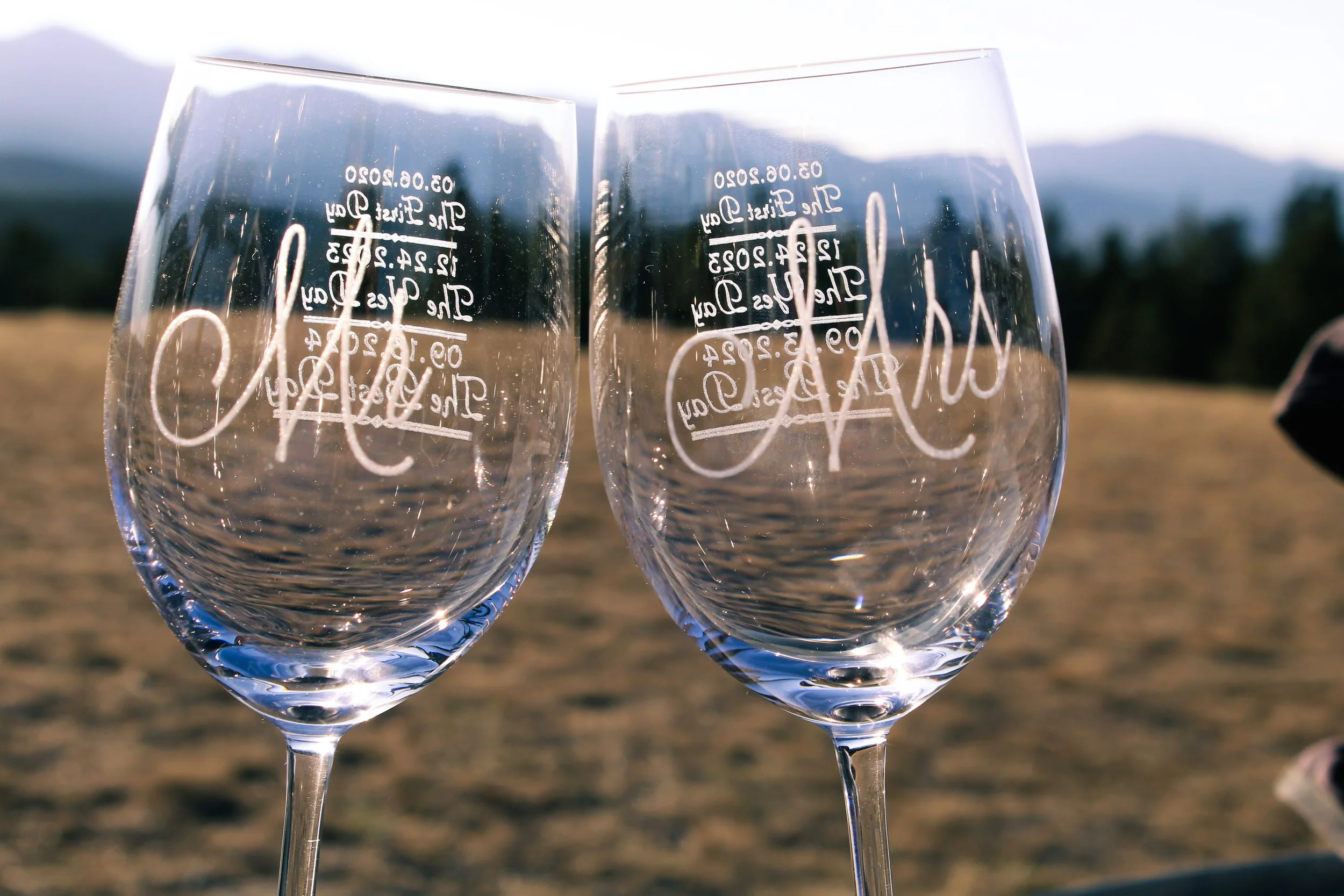 Two clear wine glasses with handwritten text, set outdoors on a sunny day with a mountainous landscape in the background.