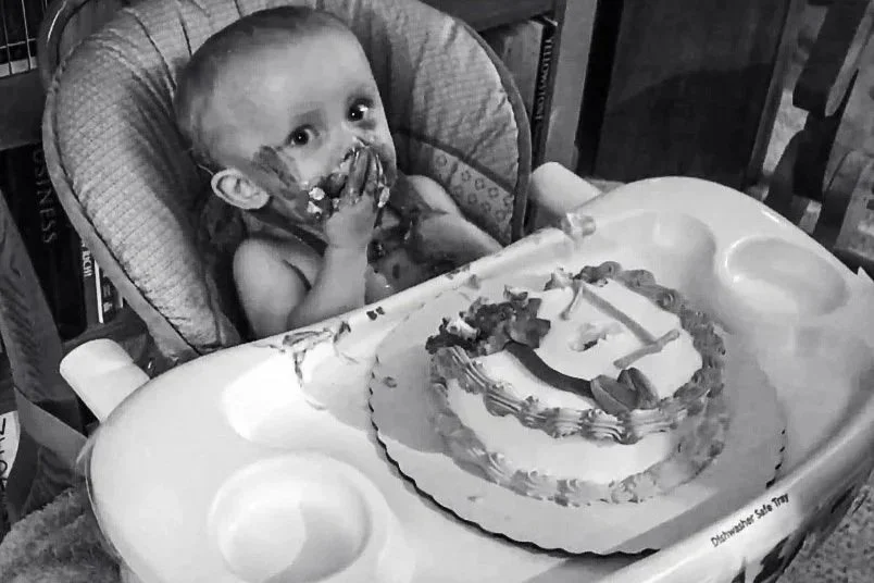 A baby sitting in a high chair with a cake in front, covered in frosting and food on their face, celebrating a birthday.