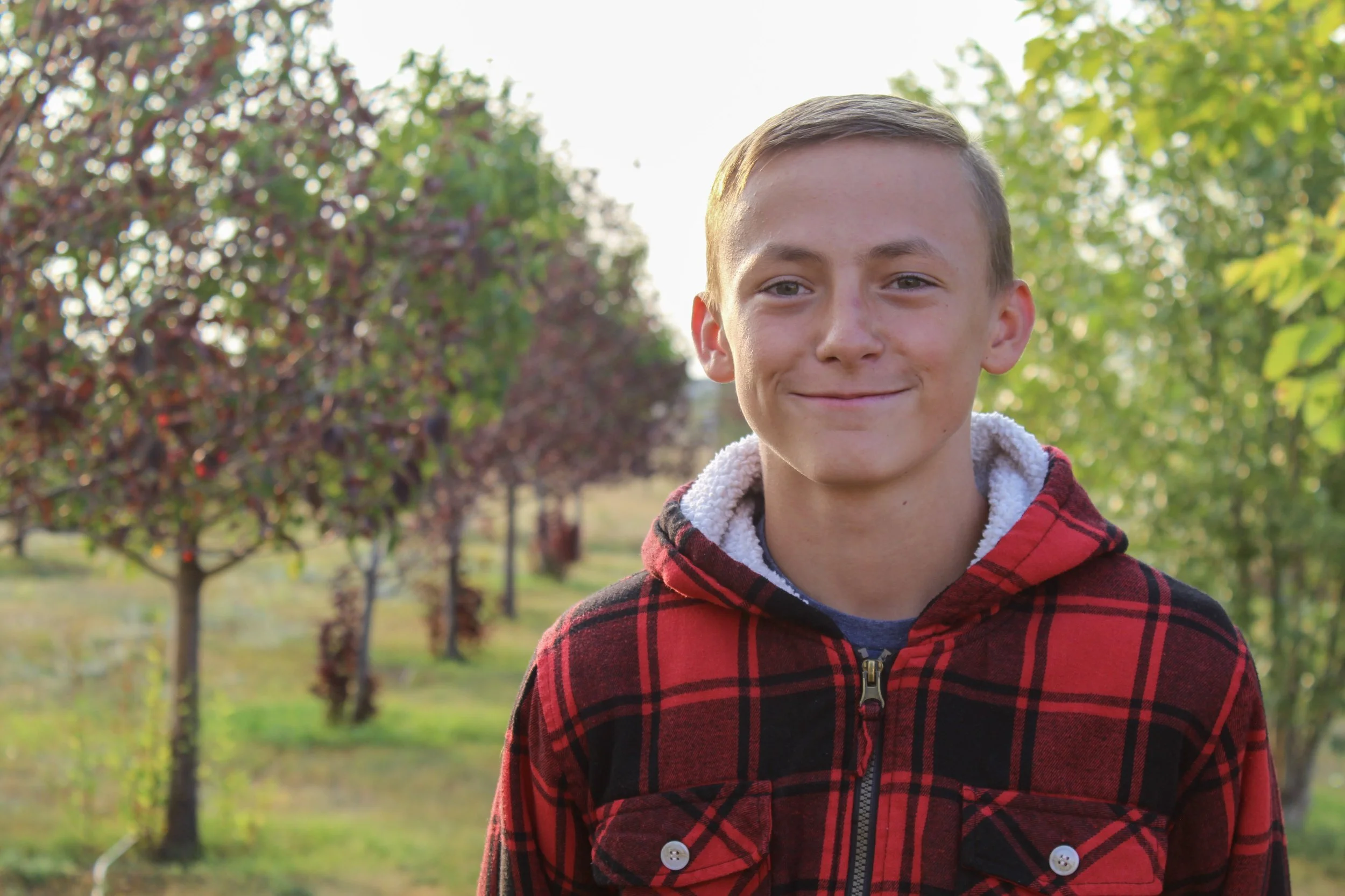 A smiling young boy with short hair wearing a red and black plaid jacket standing outdoors in a park or orchard with trees and green foliage in the background.