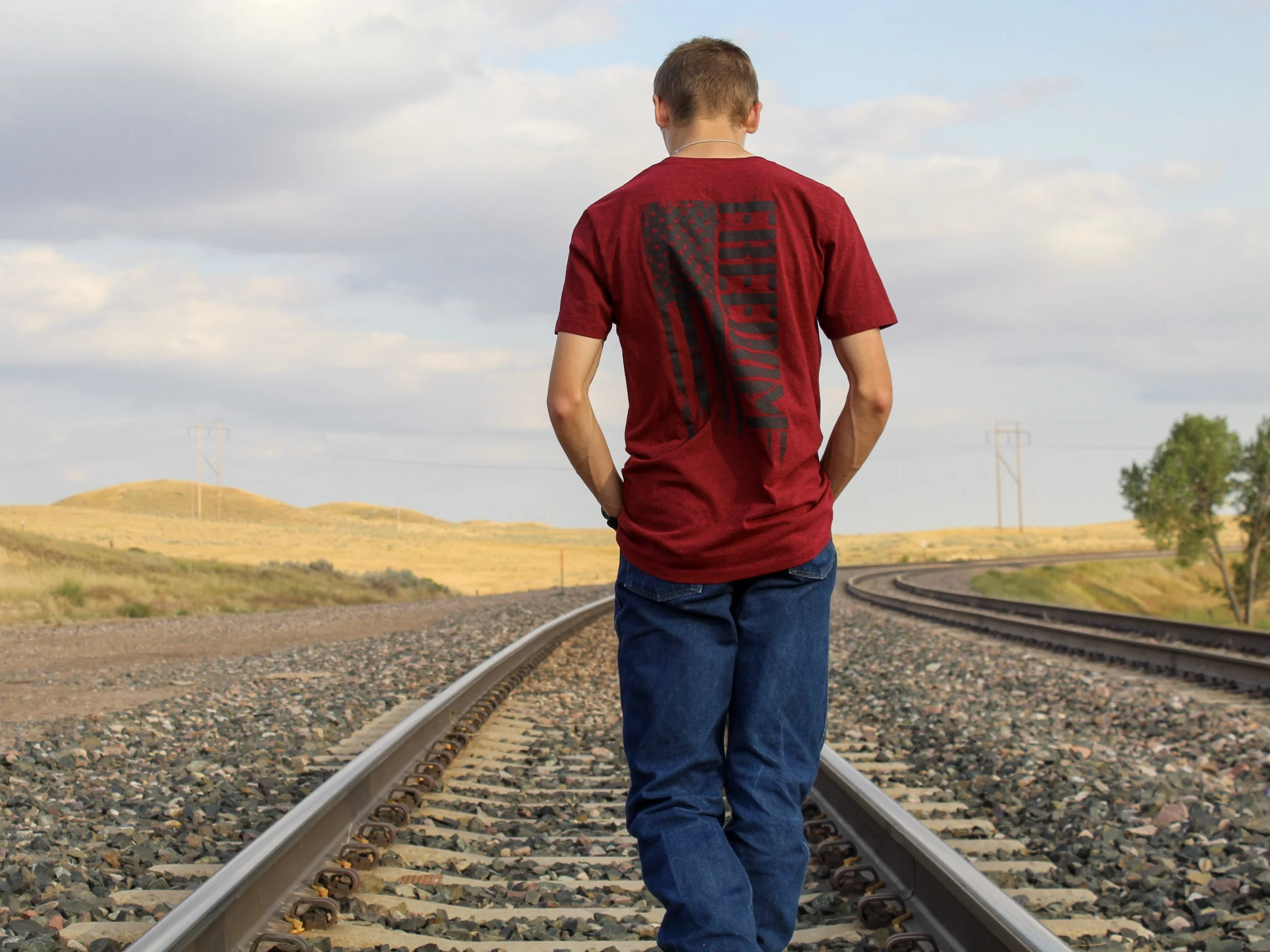 A young man walks along a set of railroad tracks in a rural landscape with rolling hills and trees, under a partly cloudy sky.