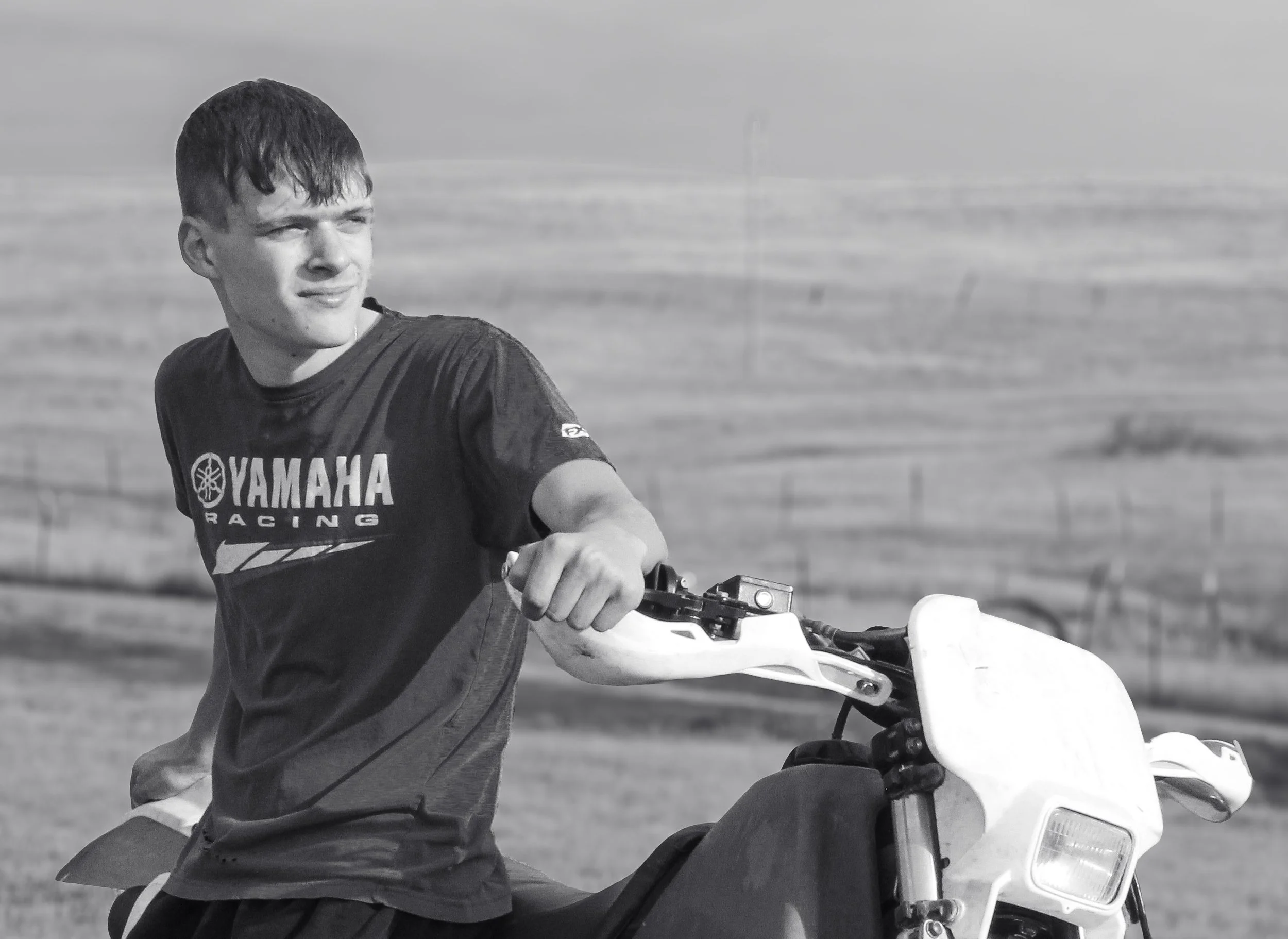 A young man in a Yamaha racing t-shirt sitting on a dirt bike in an open field.