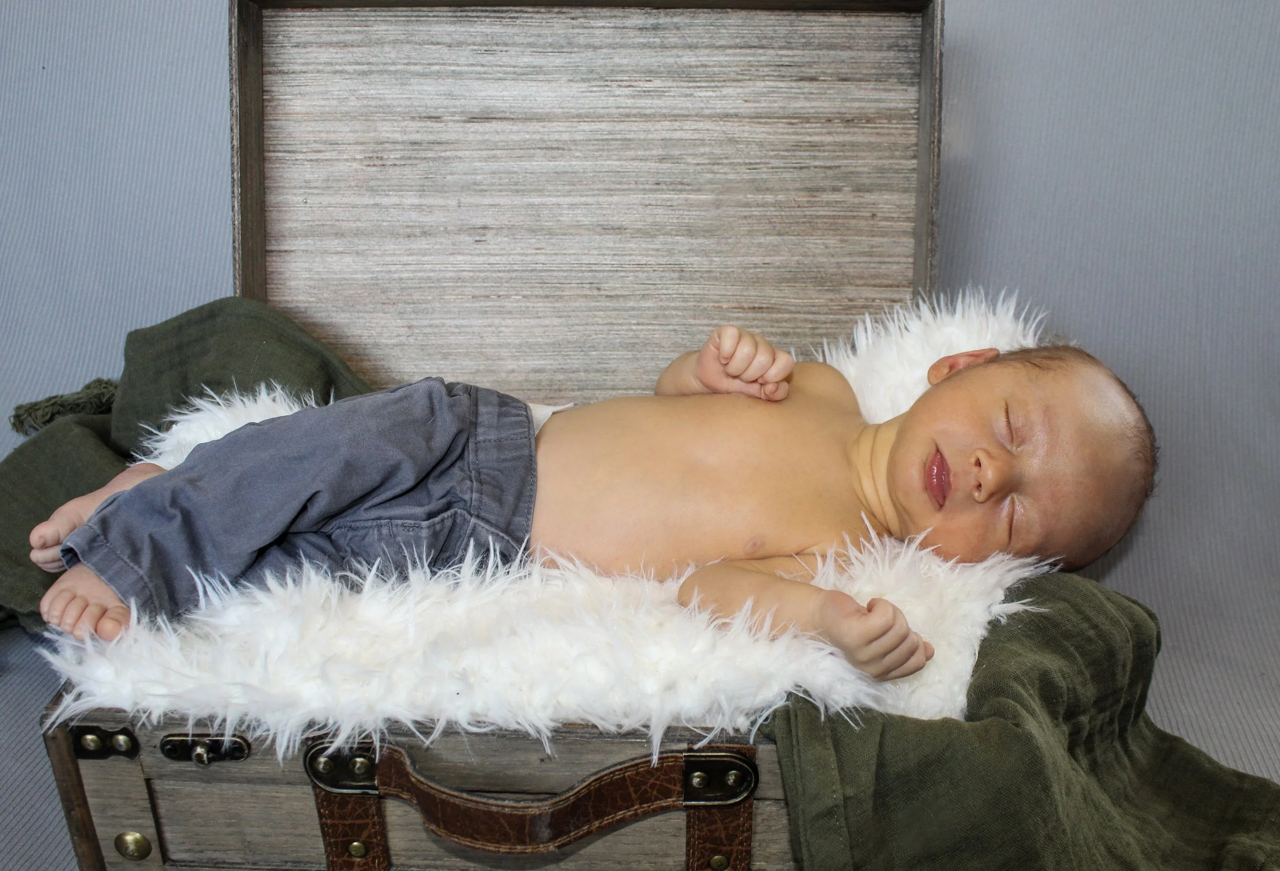 A newborn baby sleeping on a fluffy white blanket inside an open vintage-style suitcase with an olive green cloth underneath. The baby is shirtless, wearing gray pants.