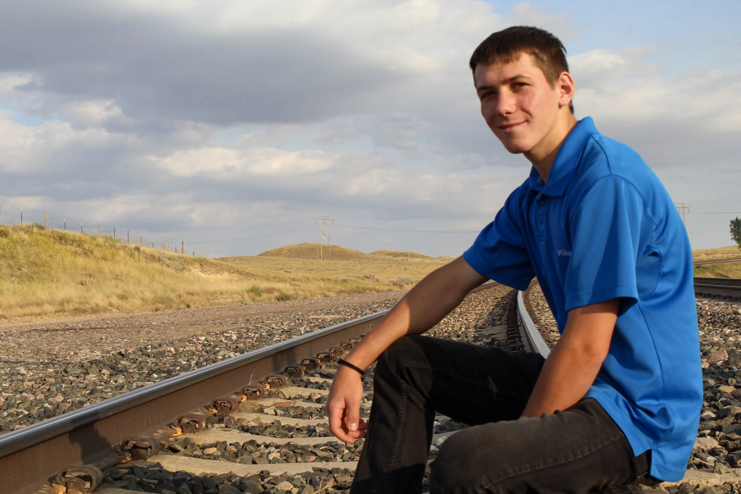 A young man in a blue polo shirt sitting on train tracks in a rural landscape with grassy hills and cloudy sky.