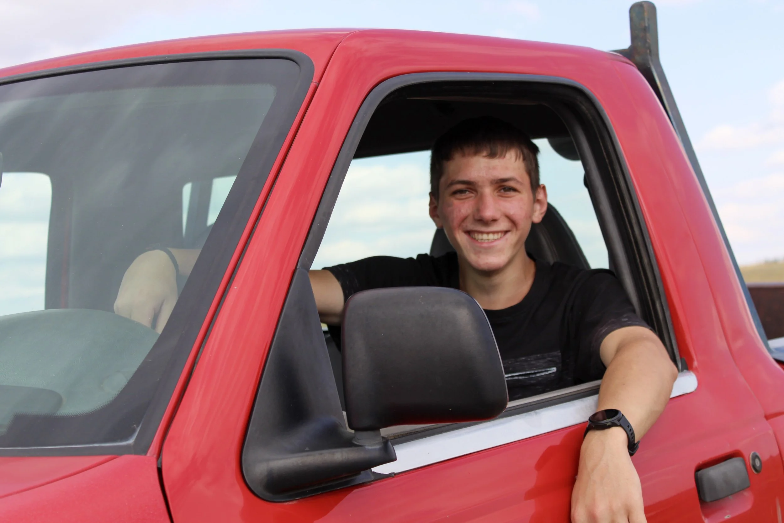 A young man with short dark hair sitting in the driver's seat of a red pickup truck, smiling and leaning out of the window with his arm resting on the door.