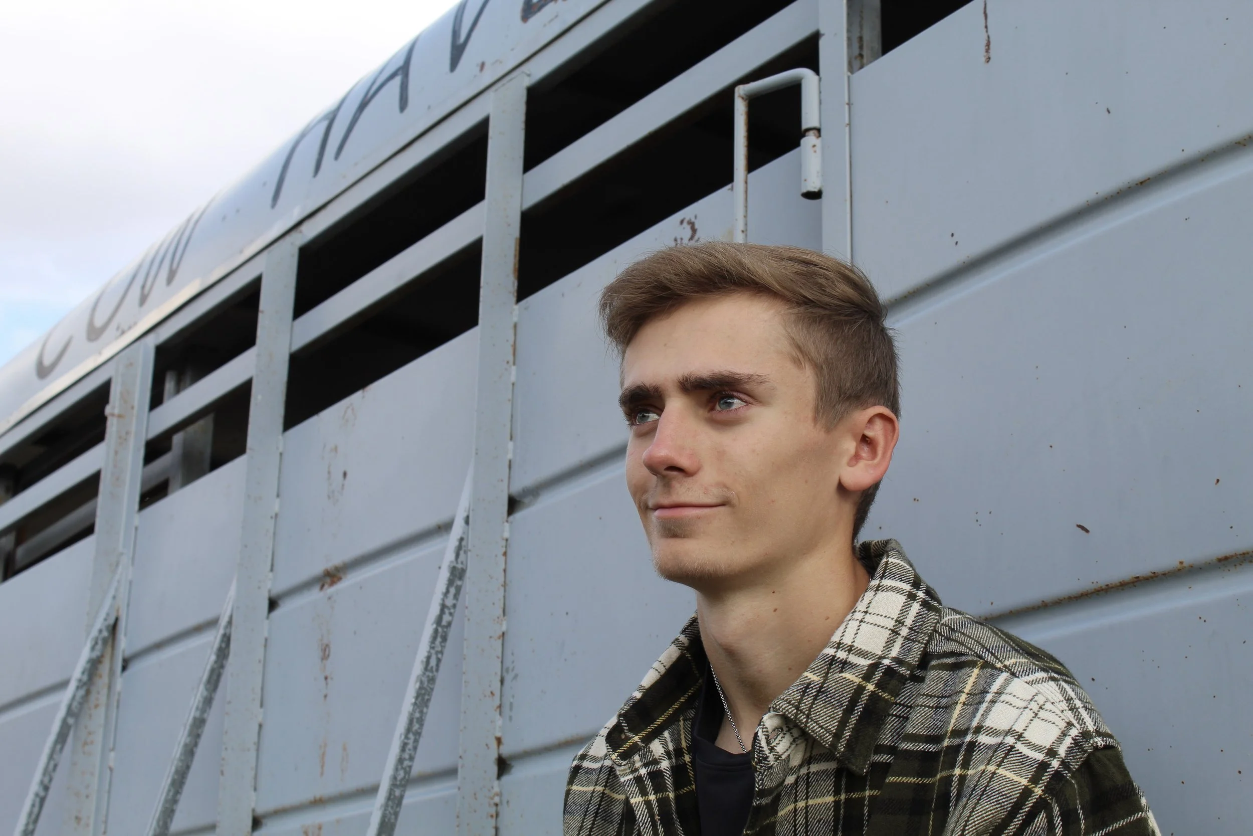 A young man with short light brown hair and blue eyes leaning against a light gray trailer, outdoors with a cloudy sky in the background.