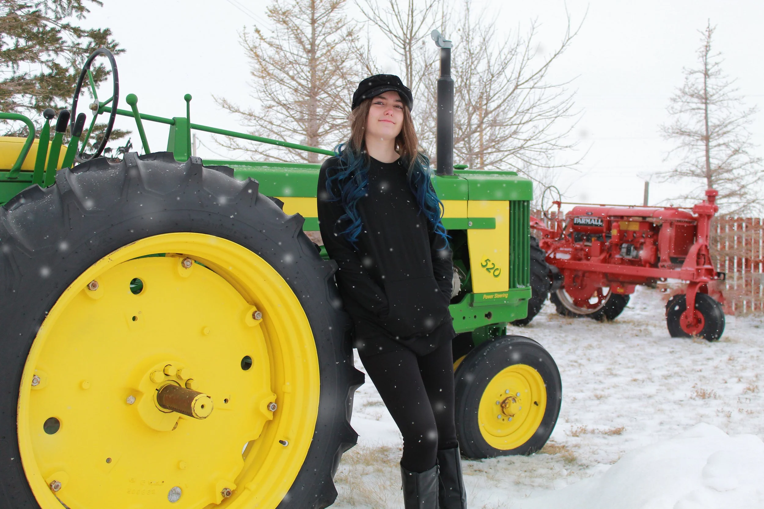 A young woman with long blue and brown hair standing outdoors in a snowy area, leaning against a large green and yellow tractor.