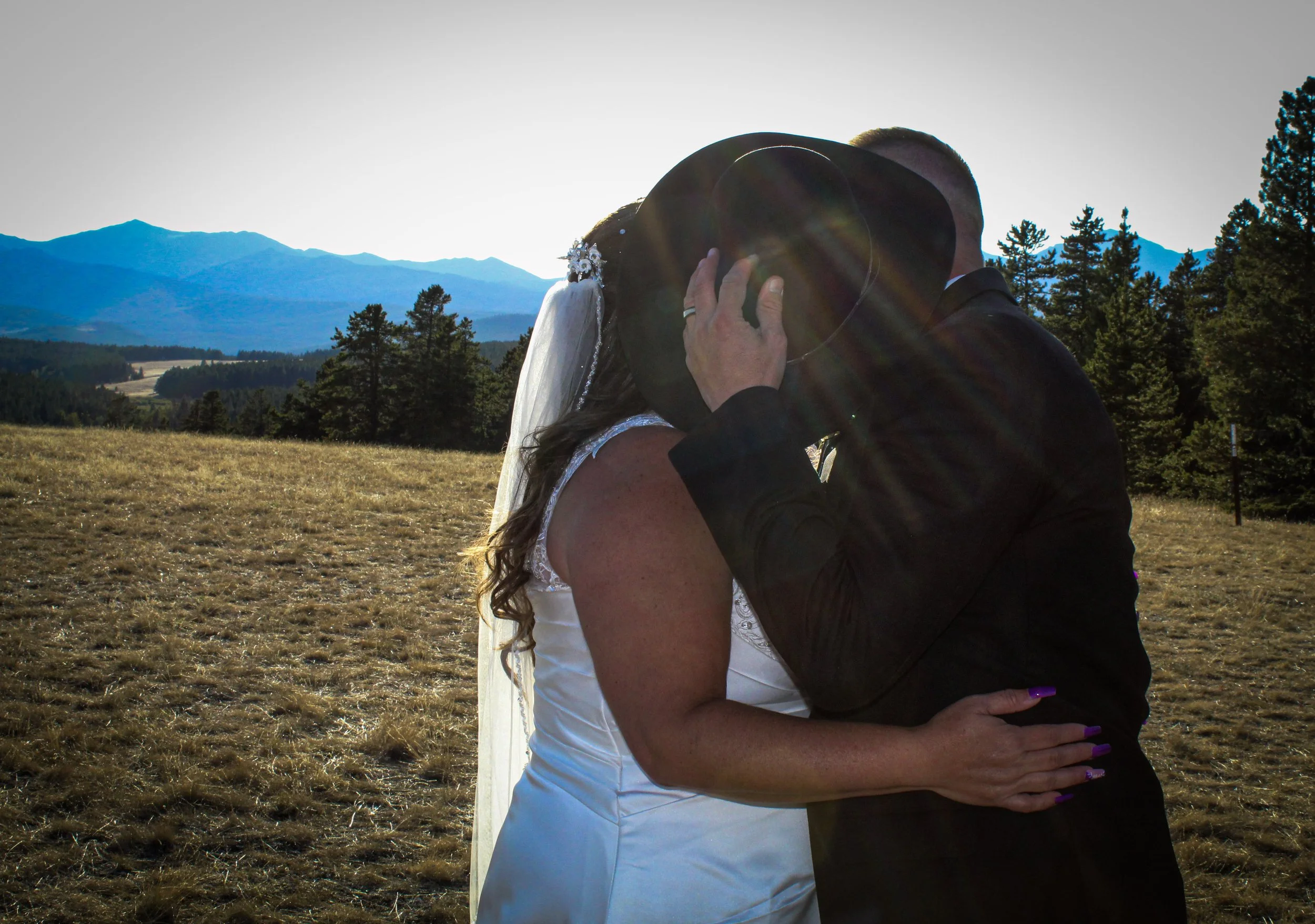 A bride and groom share a kiss outdoors at sunset, with mountains and trees in the background.