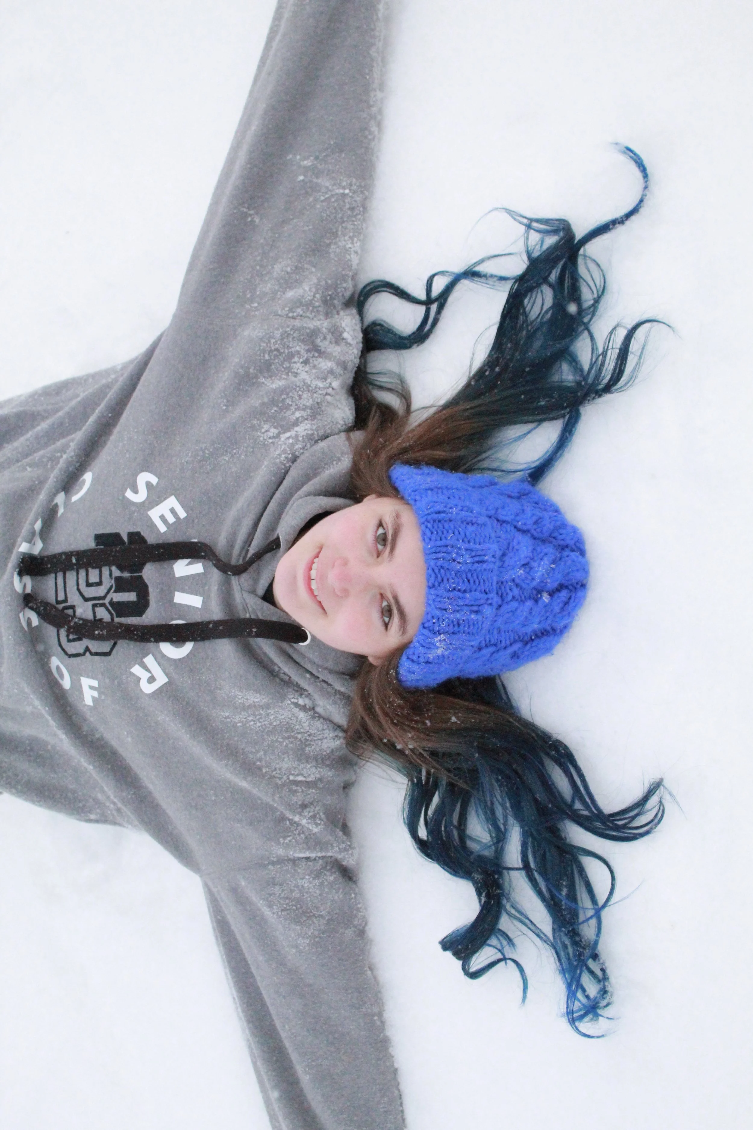 Young woman lying in snow with arms outstretched, wearing a blue knit hat and gray hoodie.