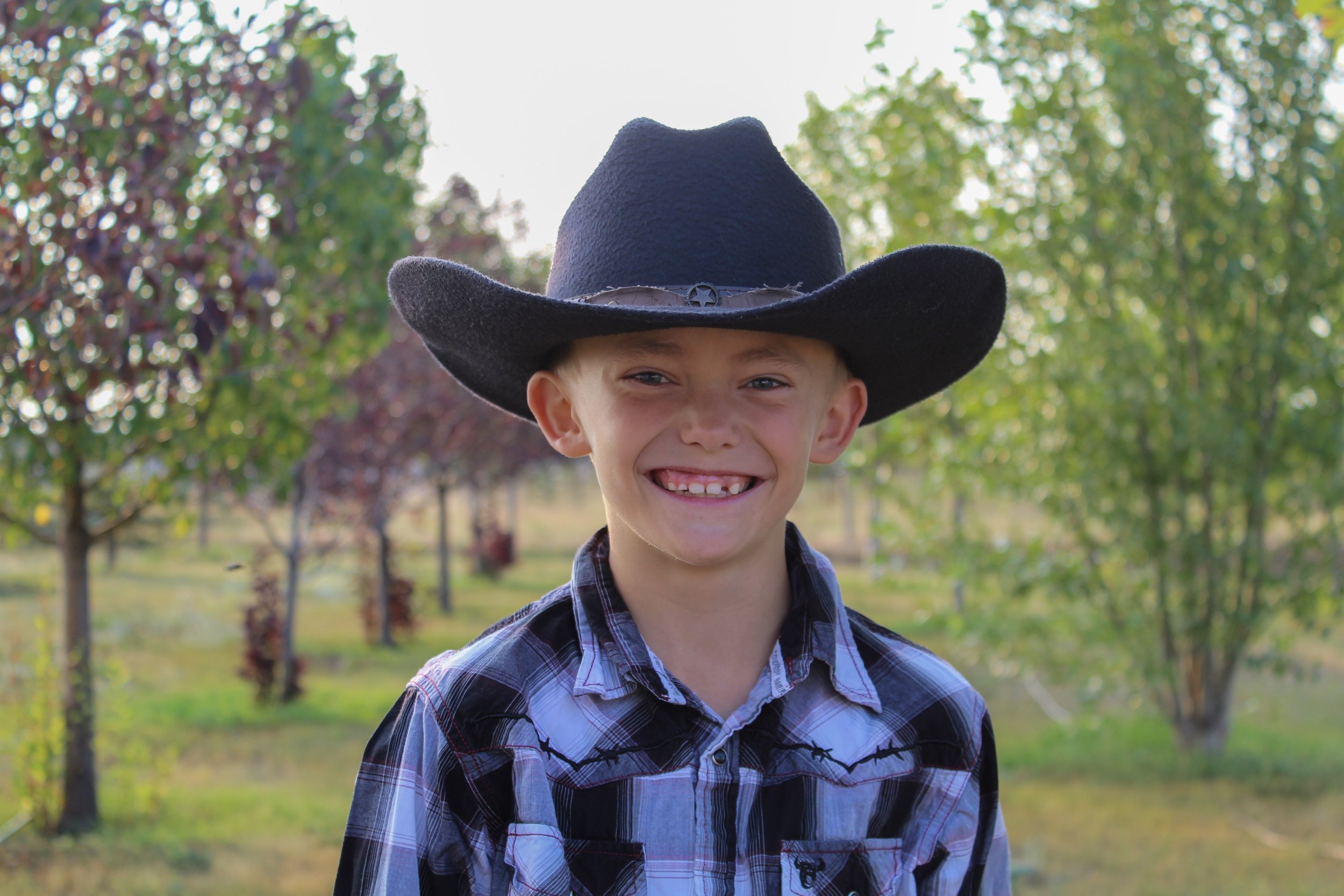 A smiling boy wearing a black cowboy hat and plaid shirt standing outdoors with trees in the background.