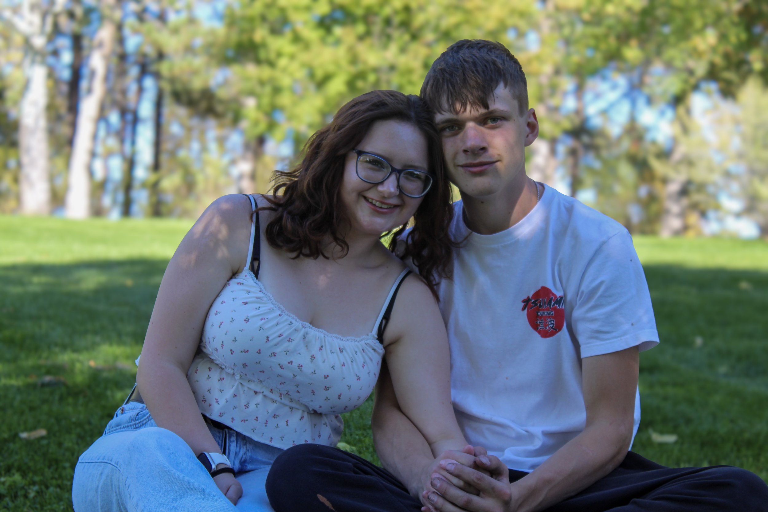 A young couple sitting on grass in a park, smiling, with trees in the background.
