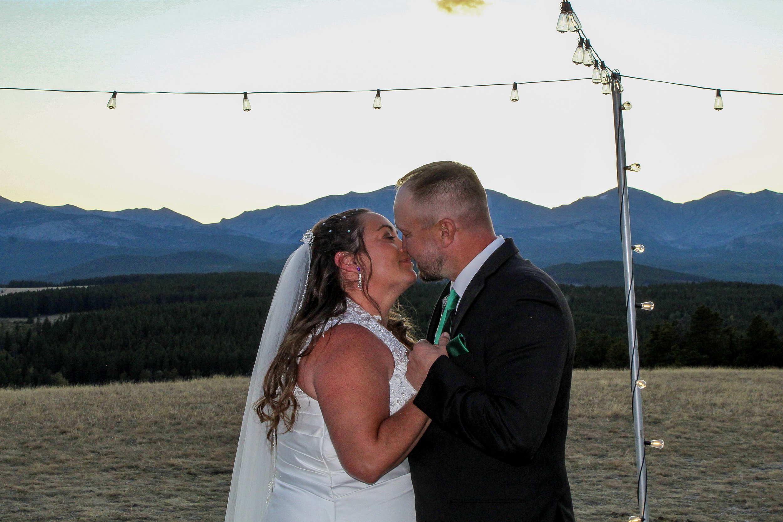 A bride and groom share a kiss during their outdoor wedding at sunset, with mountains and string lights in the background.