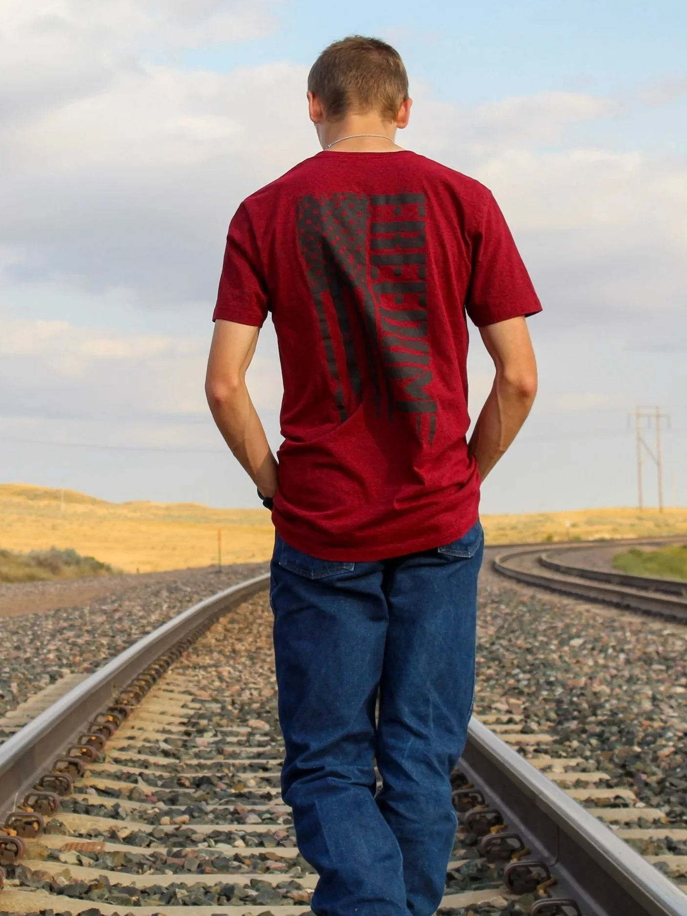 A person with short hair wearing a red T-shirt with black graphic design on the back, blue jeans, standing on railway tracks in a rural landscape.