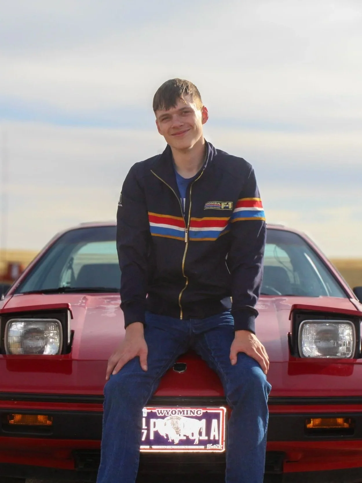 Young man sitting on the hood of a red sports car, smiling, outdoors on a cloudy day.