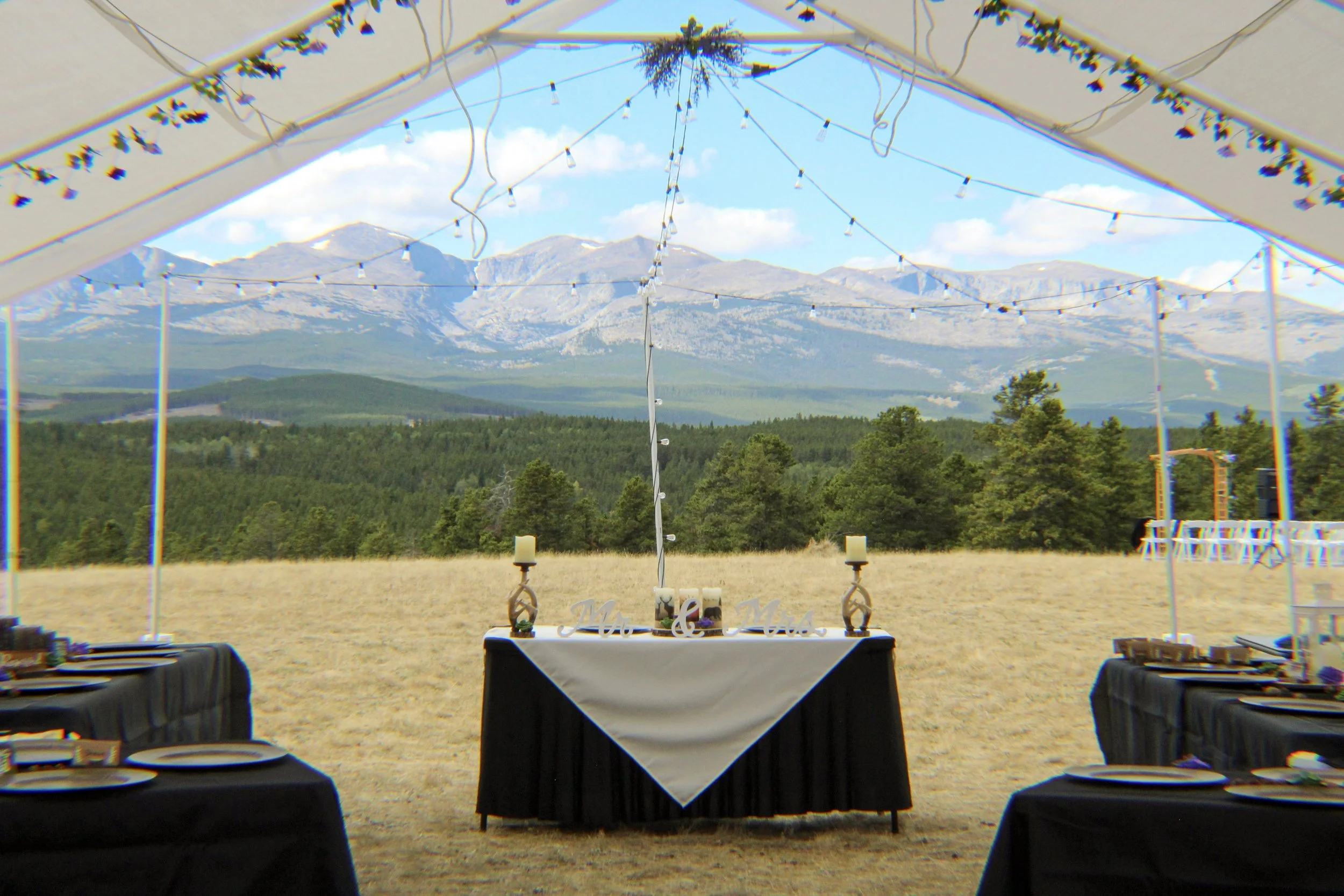 Outdoor event setup with tables, candles, and string lights, overlooking a mountain range and green forest background.