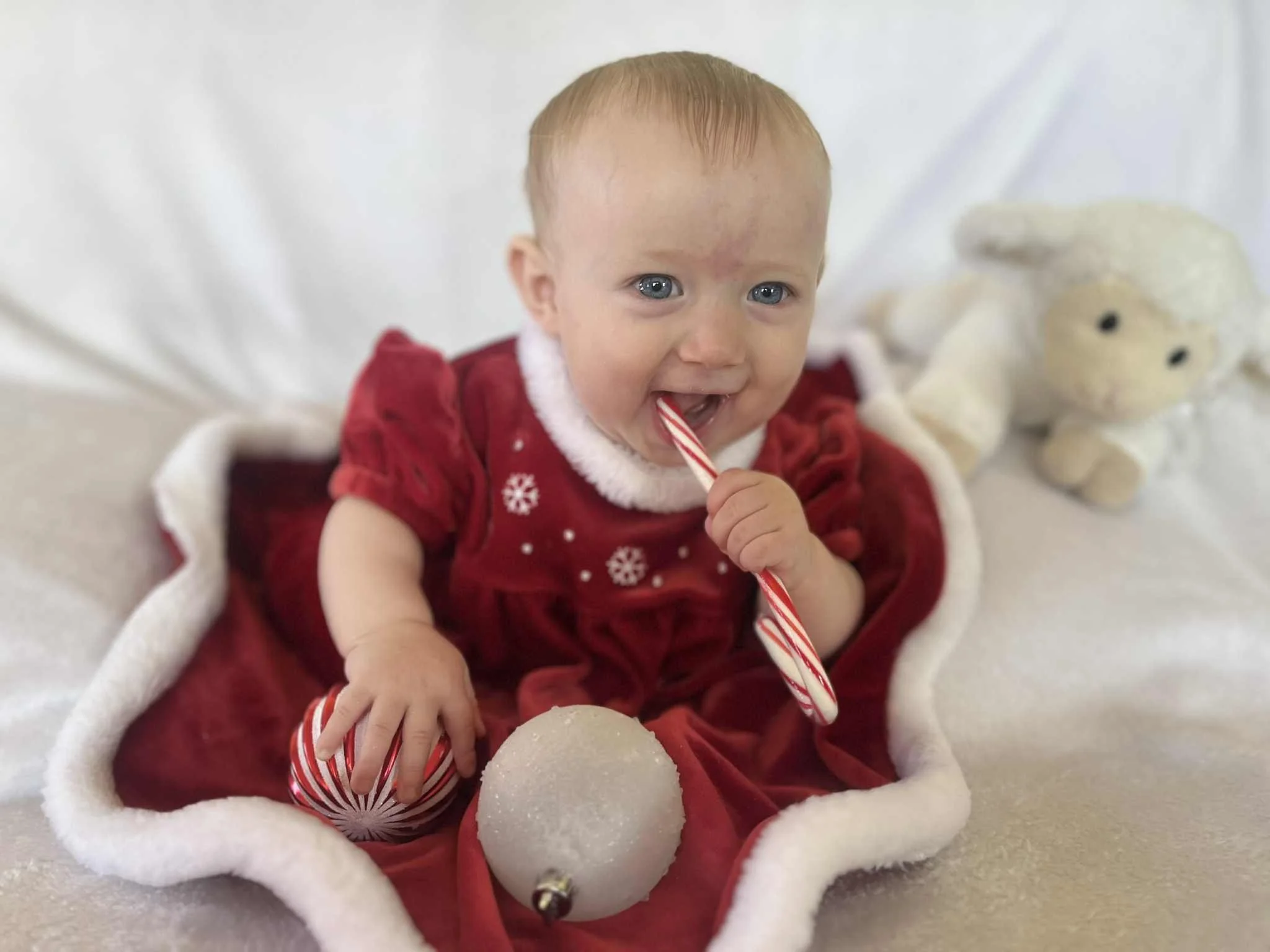 A baby in a red Christmas outfit holding a candy cane and a striped ornament, sitting on a white blanket with a plush reindeer in the background.