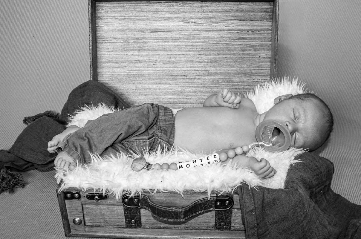 A newborn baby lying on a fluffy blanket inside a vintage wooden box with metal accents, wearing jeans, with a pacifier in its mouth and a string of beads with letter blocks.
