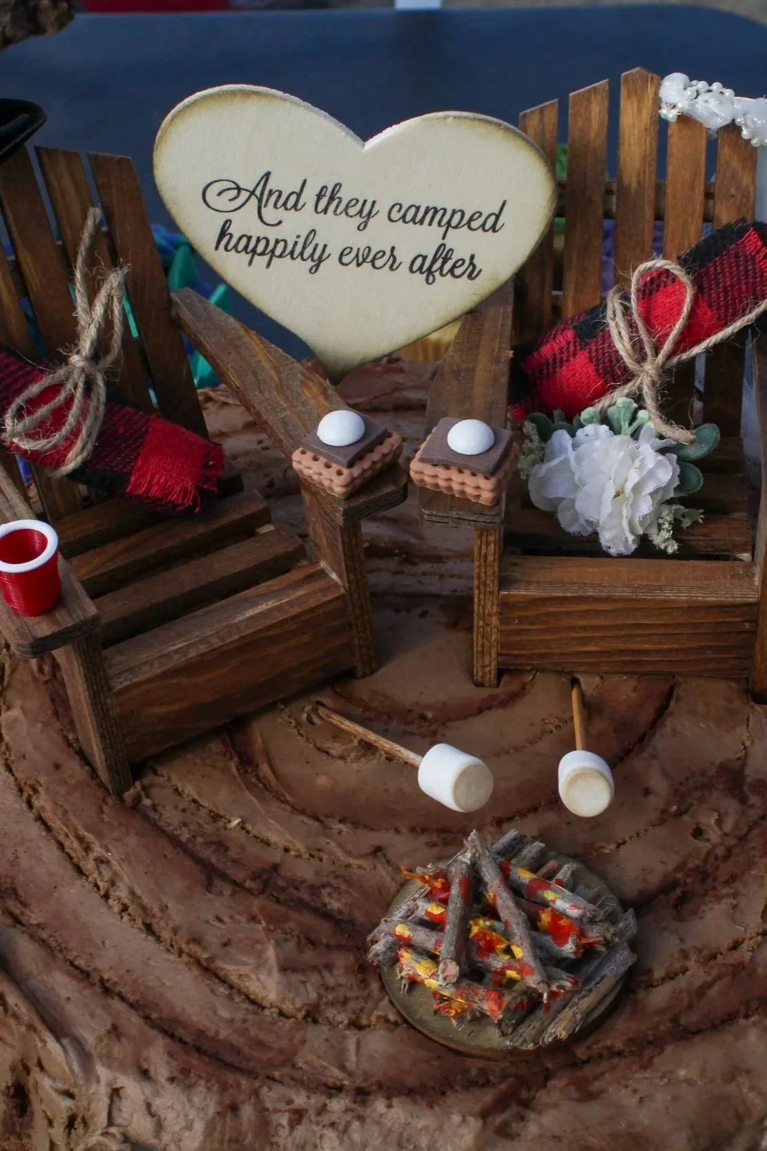 Miniature campfire scene with two wooden chairs, a heart-shaped sign reading 'And they camped happily ever after,' marshmallow roasting sticks, small faux marshmallows, a red plaid blanket, and decorative flowers on a tree trunk surface.