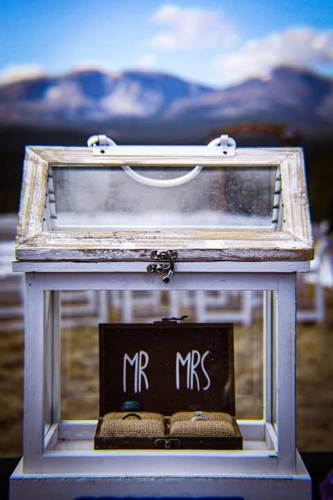 A glass display box with a wooden frame and a latch, showing wedding rings inside, with a sign reading 'MR MRS' below and outdoor chairs in the background.