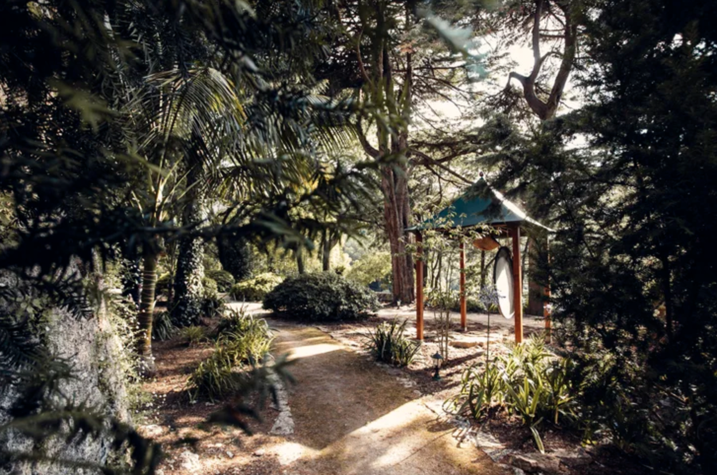 Jardin zen avec un chemin en terre, un petit temple en bois avec une cloche, entouré de plantes et d'arbres