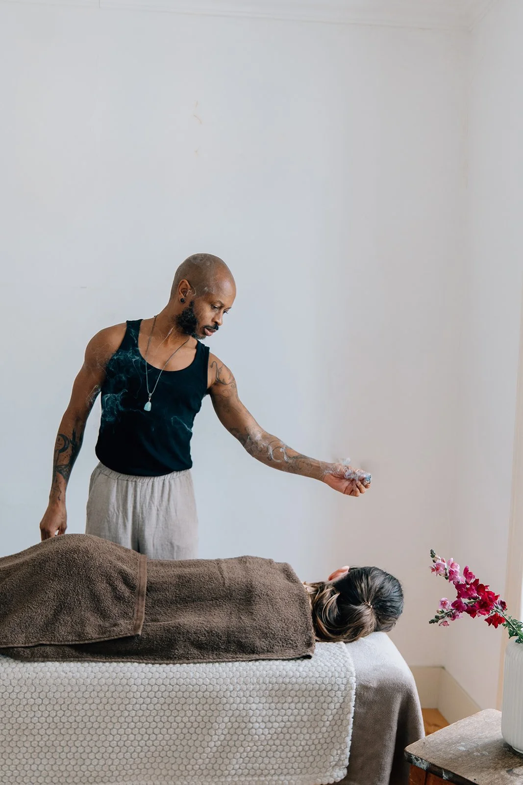 Un homme tatoué en débardeur noir donne un massage à une personne allongée sur une table, recouverte d'une serviette marron, dans une pièce calme.