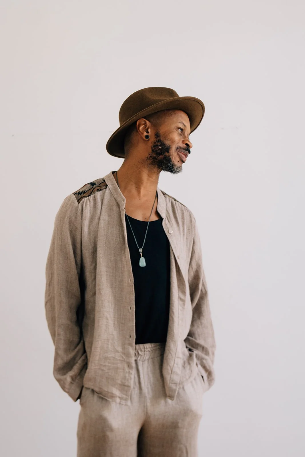 Homme portant un chapeau marron, une chemise beige, un t-shirt noir, et un collier avec une pierre turquoise, posant de profil contre un fond blanc.