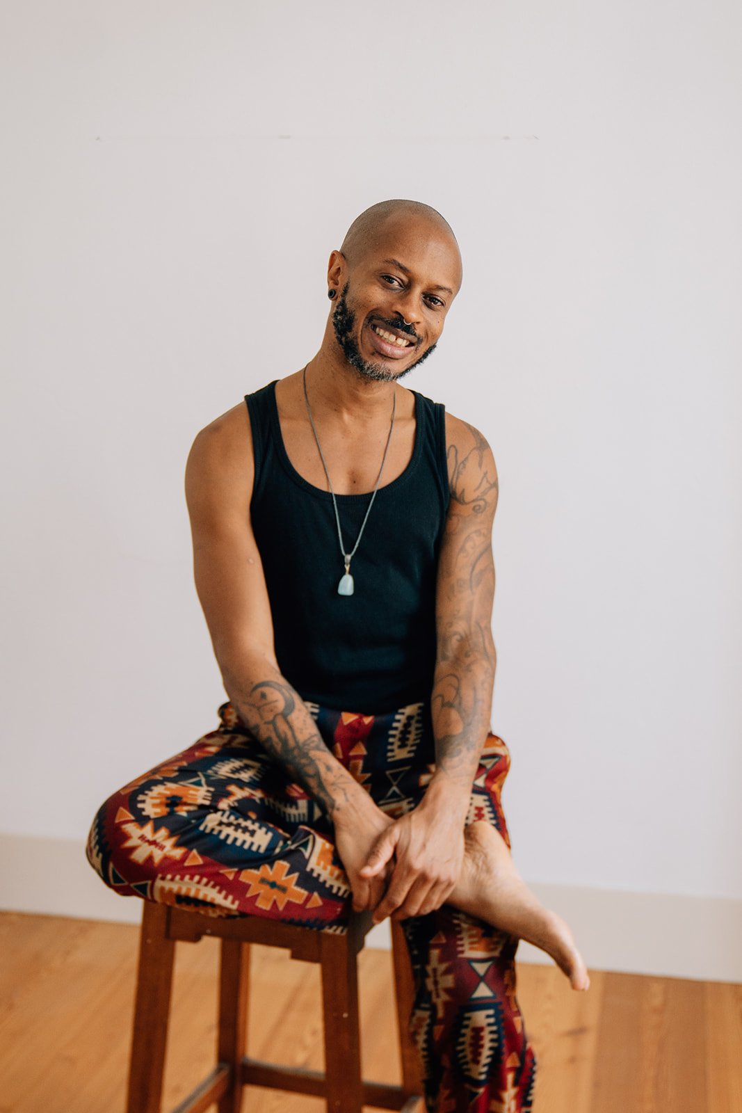 Homme à la peau sombre, chauve, avec une barbe, portant un t-shirt sans manches noir et un pantalon aux motifs géométriques, assis sur une chaise en bois, souriant à la caméra, avec un mur blanc en arrière-plan.