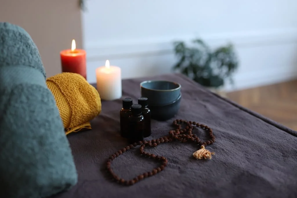 Bougies allumées, rouleaux d'essuie-tout, bouteilles d'huile essentielle, bol, mala de prière, et un tapis de massage ou de yoga sur une table en bois, dans une ambiance calme et relaxante.