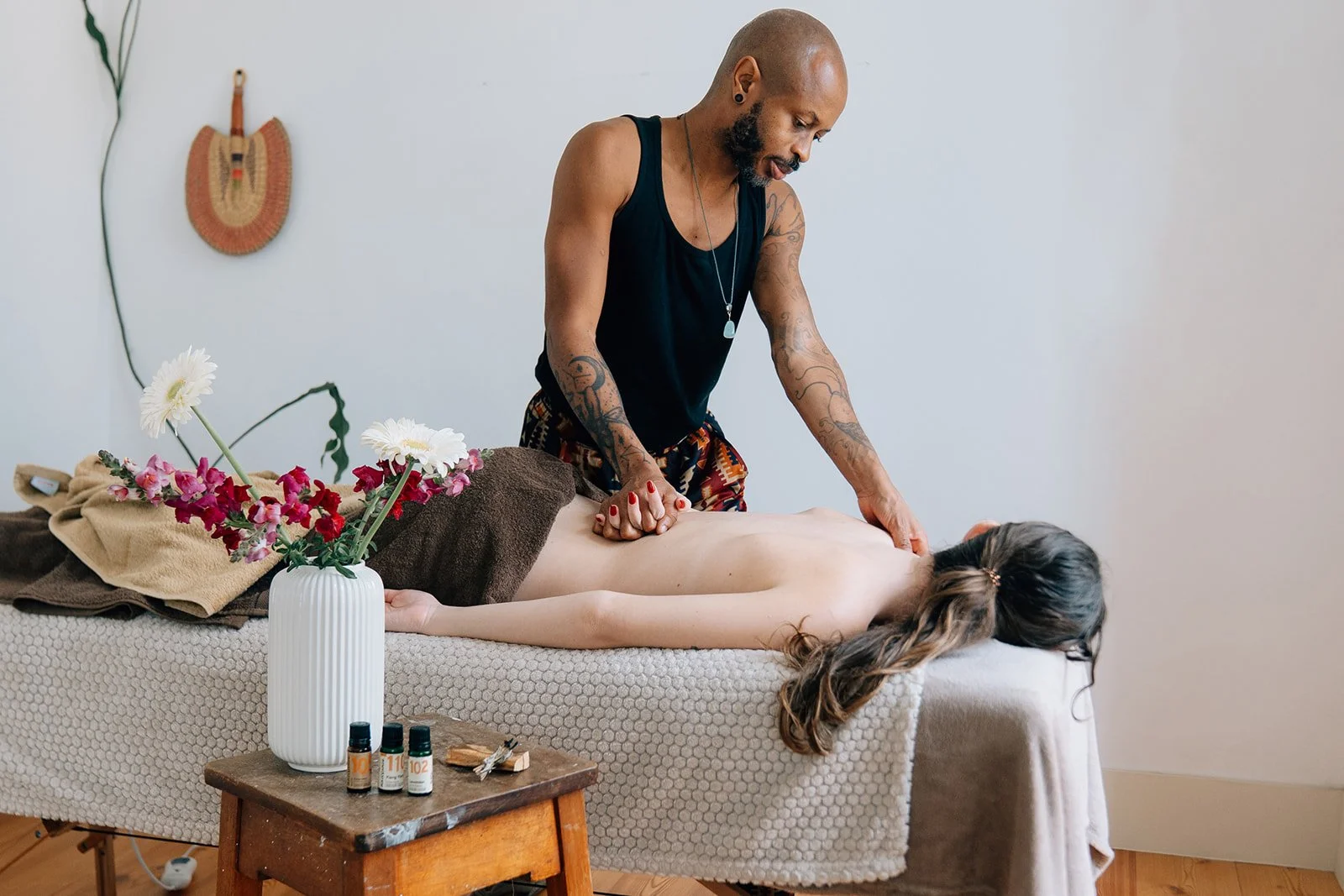 Un homme donne un massage à une femme allongée sur une table de massage, dans une pièce calme avec des fleurs dans un vase et des bouteilles d'huiles essentielles.