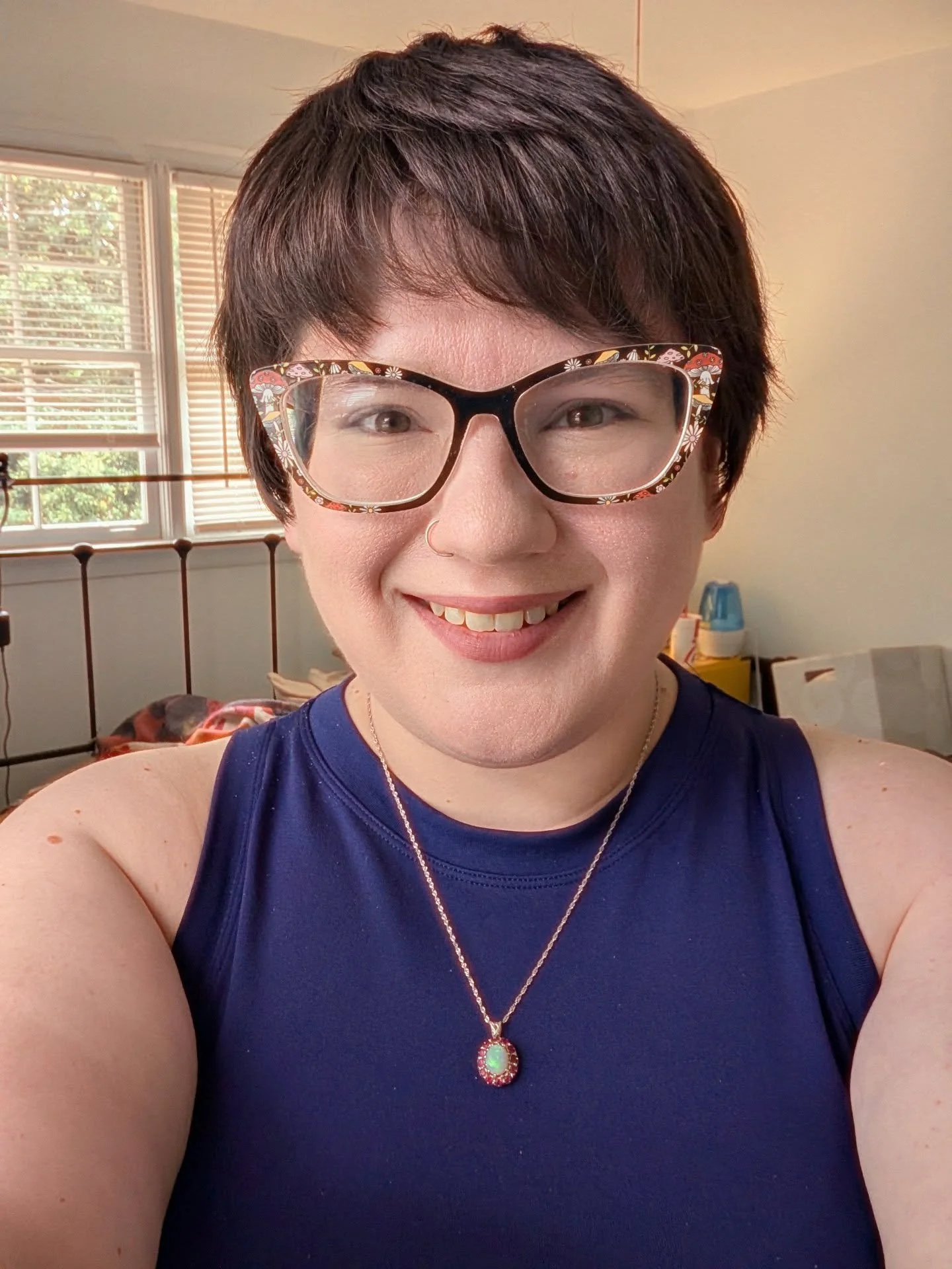 A person with short brown hair, wearing large floral-patterned glasses, a nose ring, a gold necklace with a green pendant, and a sleeveless navy blue top, smiling in a well-lit room with windows and blinds in the background.
