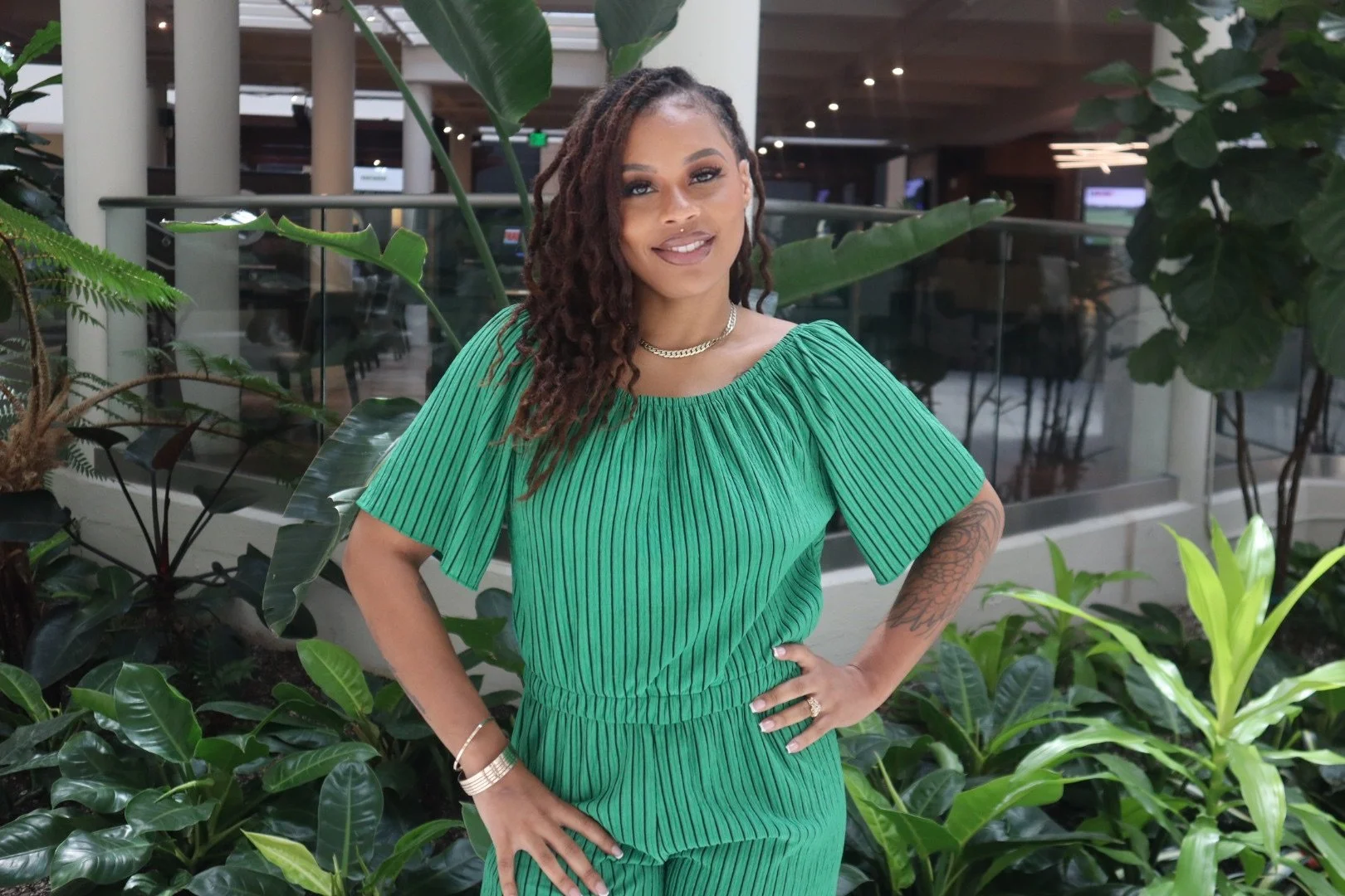 A woman with medium skin tone, brown dreadlocks, and tattoos on arms, standing with hands on hips, smiling in an indoor space with tropical plants and glass railings.